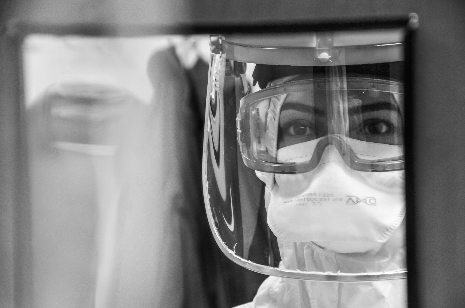 A scientist, with goggles, a paper mask, and a big plastic mask over her head, looks through a small window. New data suggests that Paxlovid rebound is uncommon (and no higher than for those not receiving the drug).