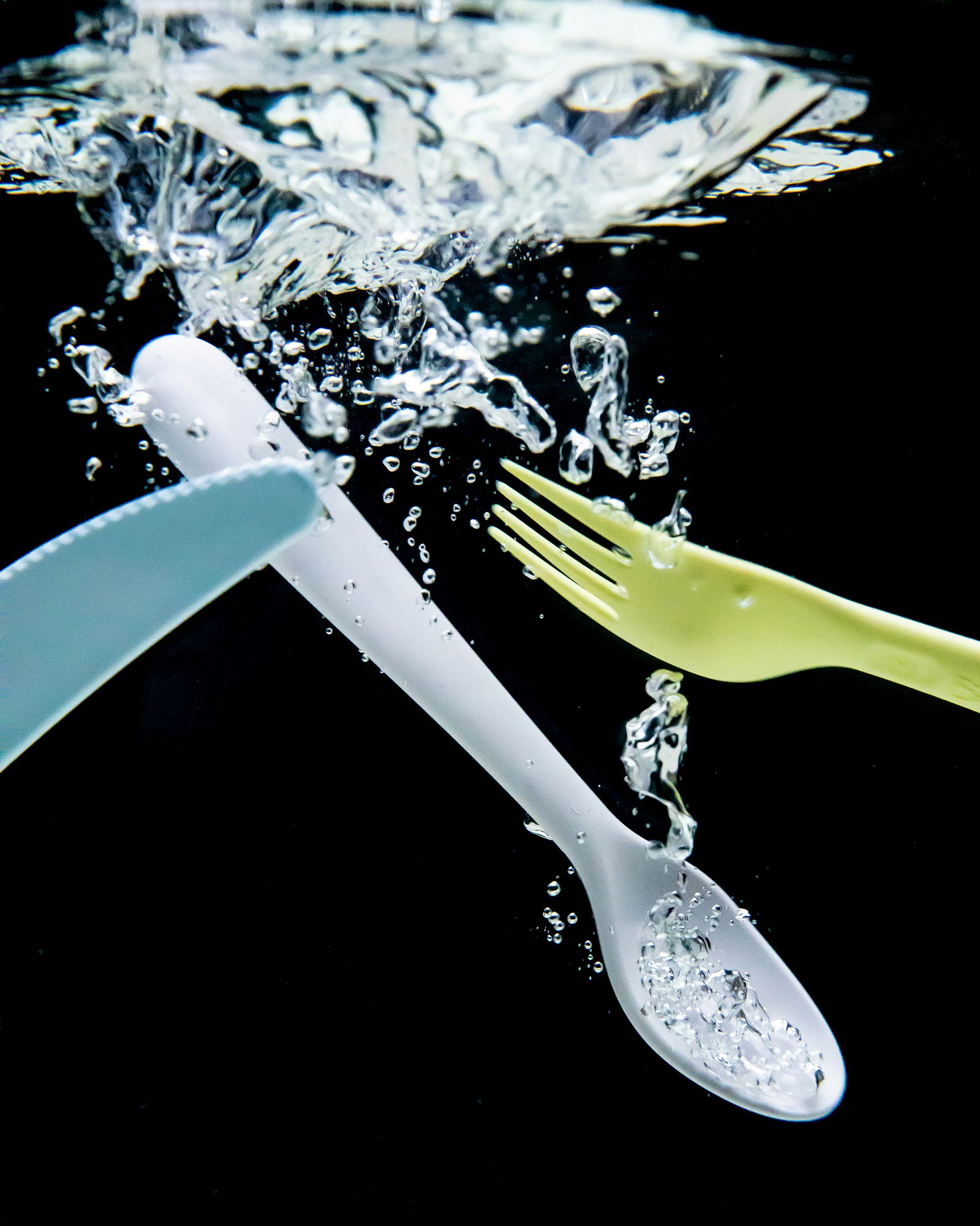 Plastic utensils —  including a blue knife, a white spoon, and a yellw fork — float in water.
