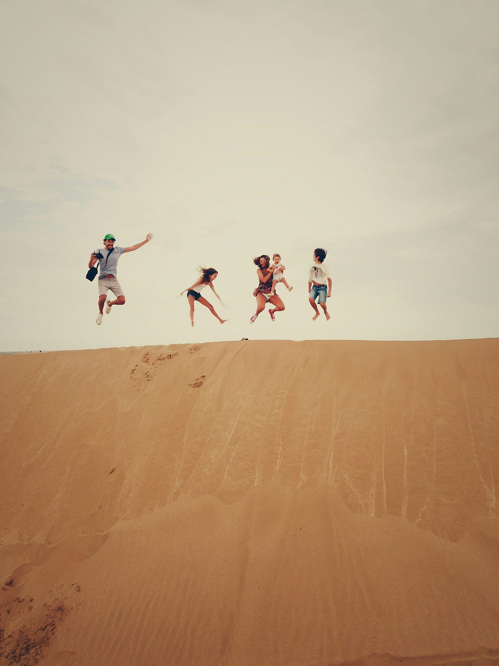 A family of five (two adults, two children, and one baby) jump on a sand dune.