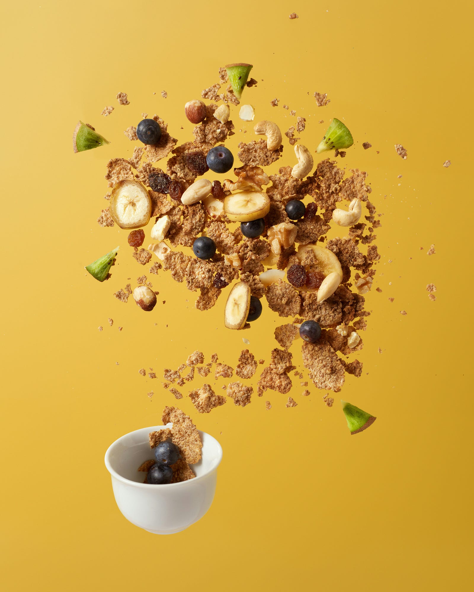 Corn flakes, banana slices, and small blueberries spring upward out of a small cup.