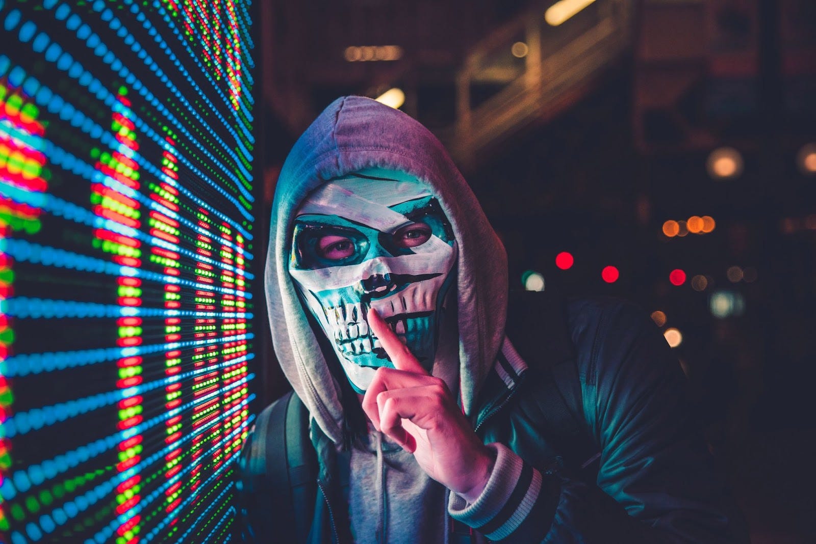 Photo of a man wearing a skull mask and a hoodie next to a digital display with a finger to his lips signaling “be quiet.”