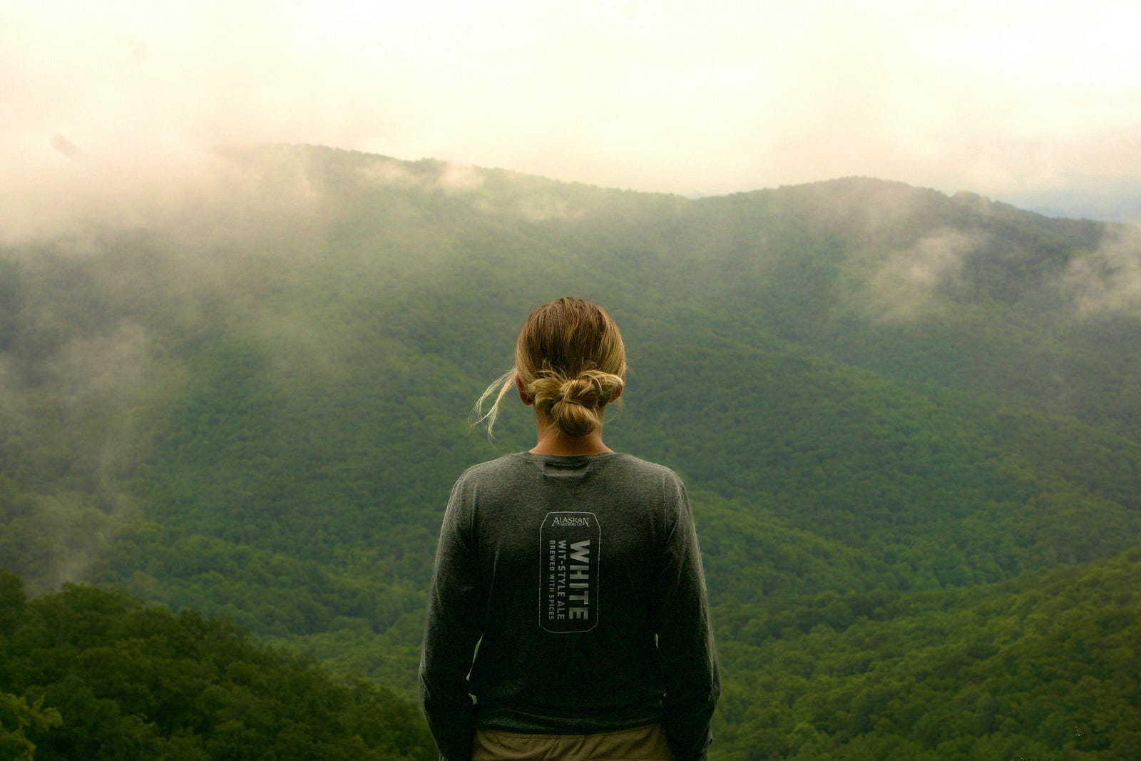 A depressed woman faces away from us as she looks over moutain valleys.