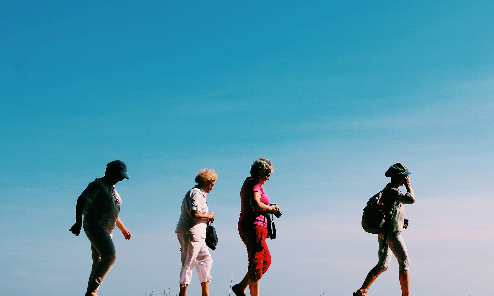 Four older individuals walk in a line from left to right, the open sky in the background. Group activities can lower fall risk.