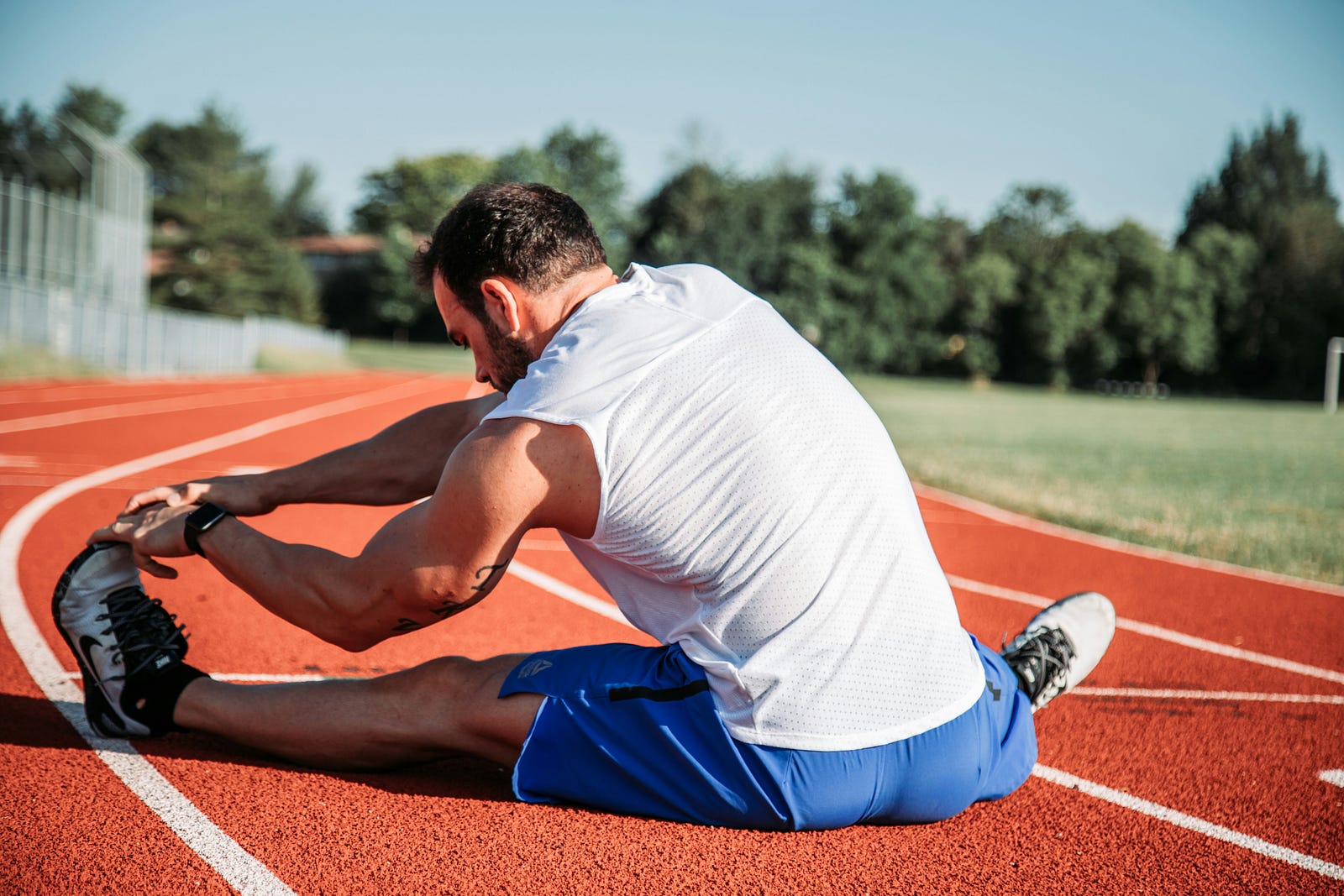 A man sits on a track as he stretches by grabbing the tip of his left shoe.