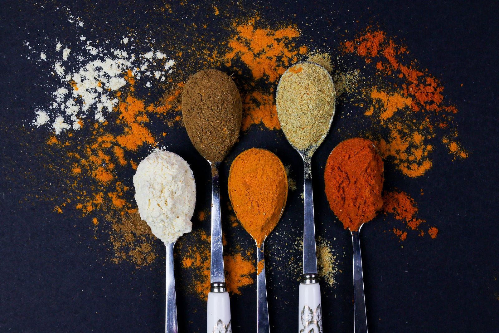 Five spoons emerge from the image bottom, with the utensils chock full of spices (colored white, brown, orange, teal, and deep red from left to right). The spices spill onto the background. Black background. Using spices can help avoid too much-added salt in our food.