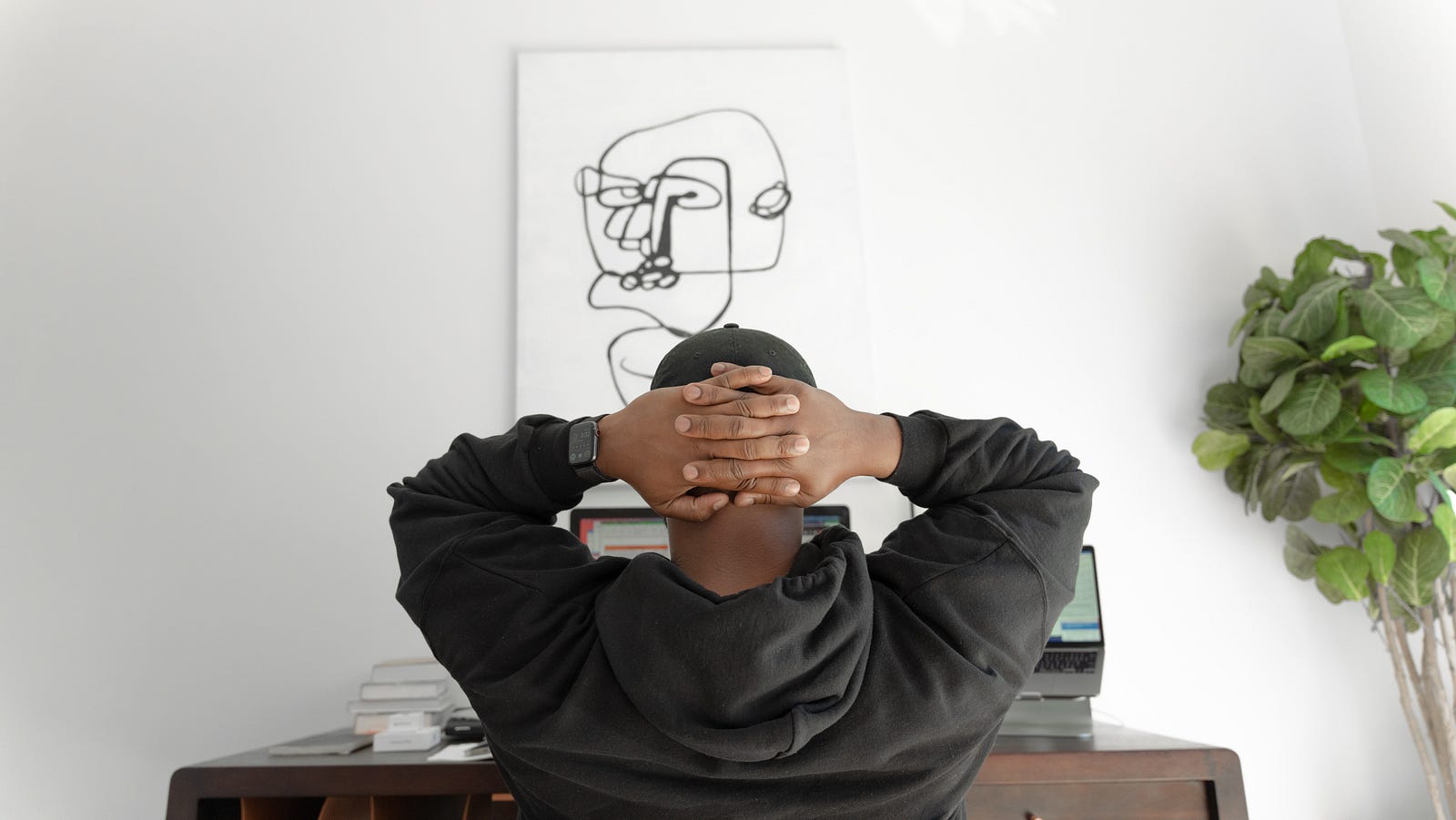 A man sits, hands behind his head, at a desk. He is focused on an abstract drawing of a head on the wall. Total sedentary time is detrimentally linked with triglycerides, waist circumference, two-hour glucose, and insulin levels.