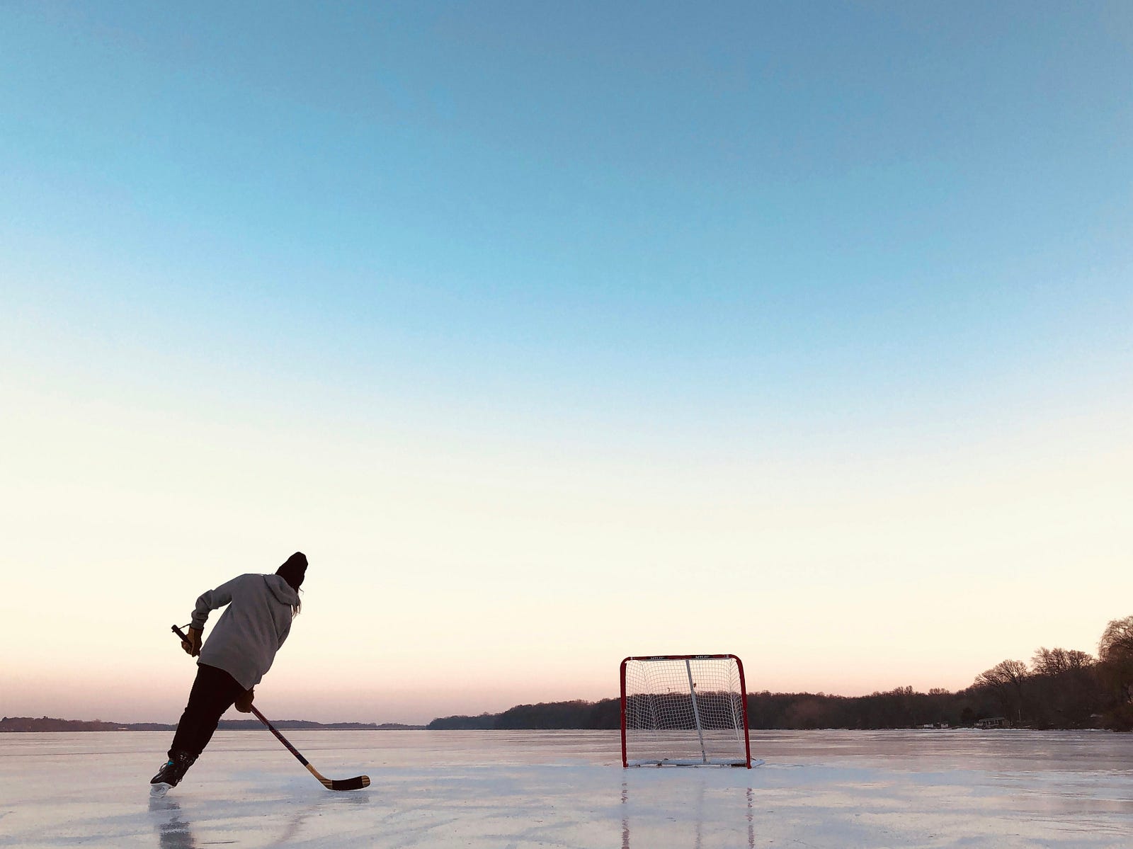 A person skates away from us on an outdoor frozen lake, hockey stick in hand. There is a net in the background.