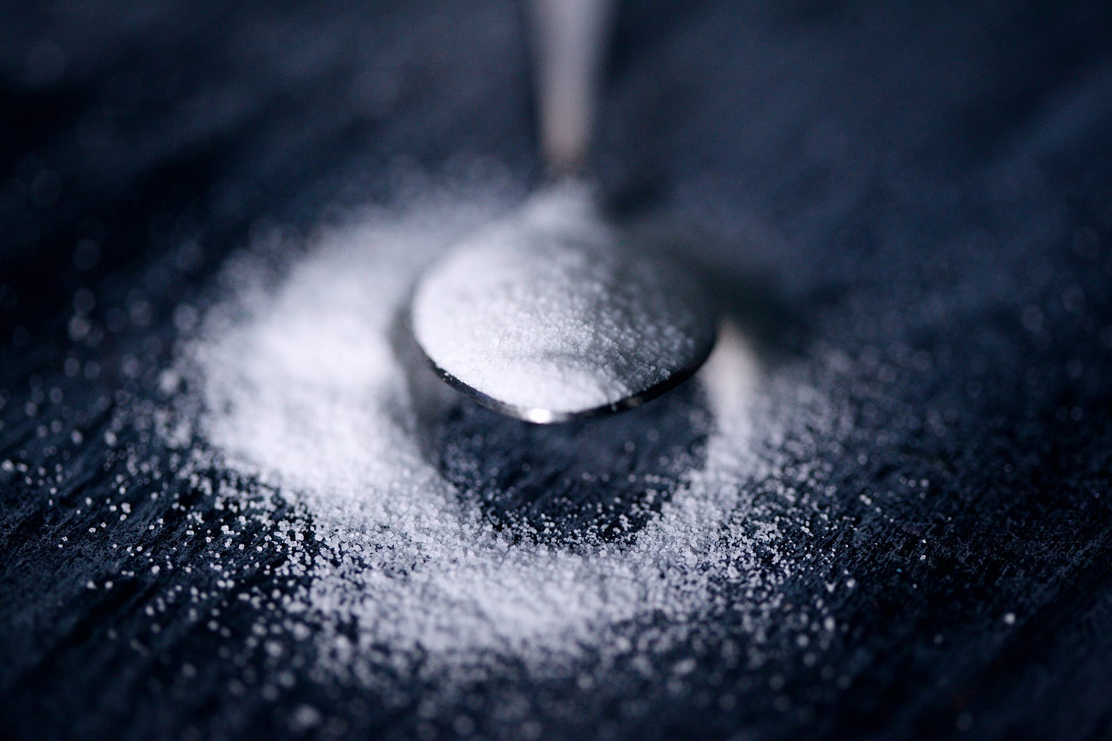 A narrow silver spoon emerges from the image top, holding sugar. More sugar is spilled onto the black surface below. Diets high in fat and sugar can cause metabolic imbalances and inflammation, disrupting the body’s natural sleep-wake cycle. Additionally, consuming these foods has been linked to increased arousal levels, making it more difficult to transition into and maintain deep sleep.