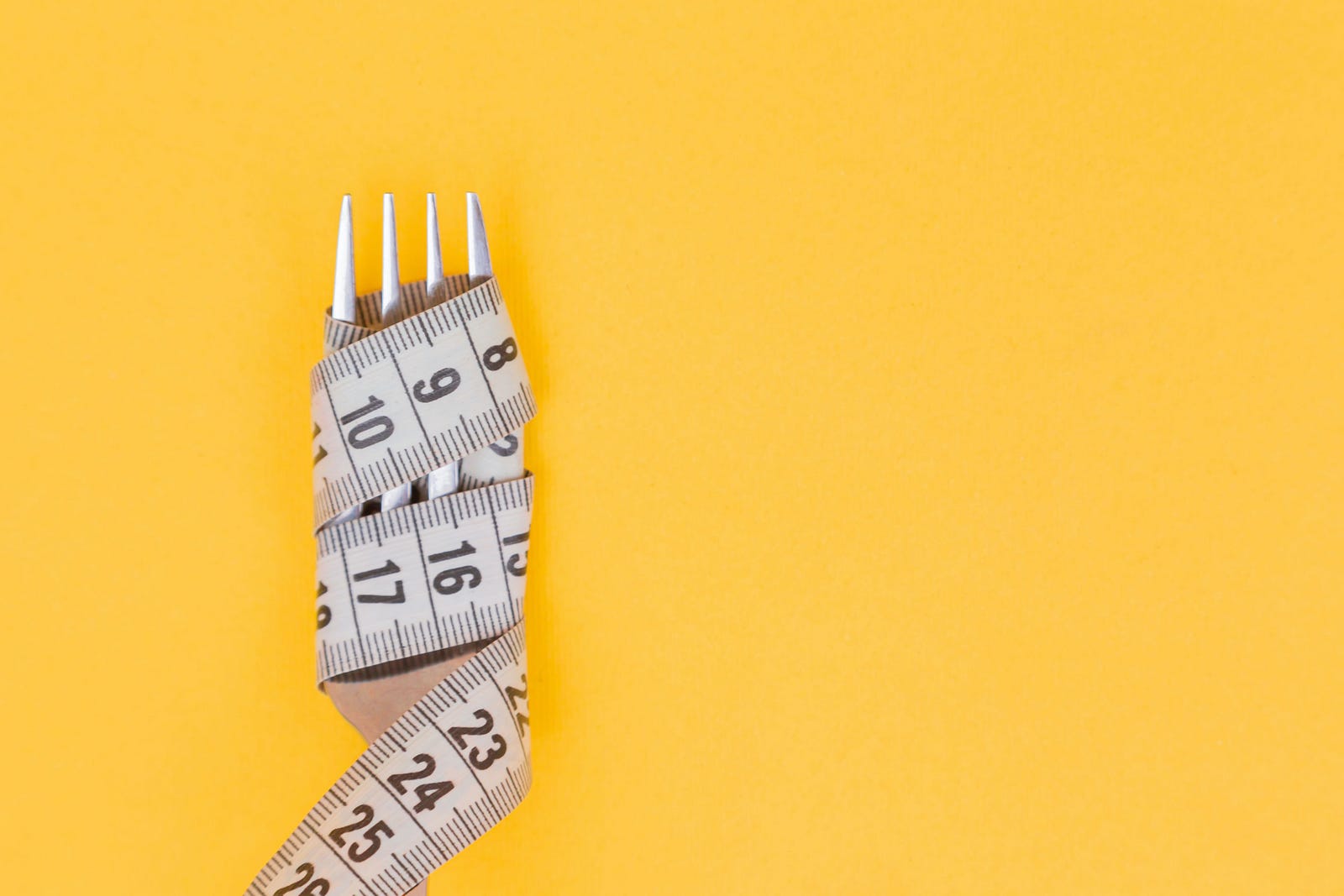 A tape measure is wrapped around the tines of a fork. Mustard-colored background. The blood-gut microbiome axis has plasticity in adults, helping to regulate food intake and keeping our weight stable. The gut microbiota creates chemical signals that determine eating behavior through interactions with the brain.