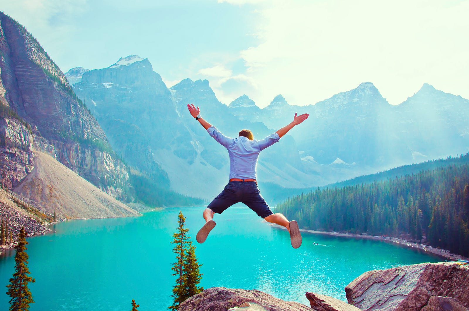 a man in blue shirt and shorts jumping against a background of lake and mountains