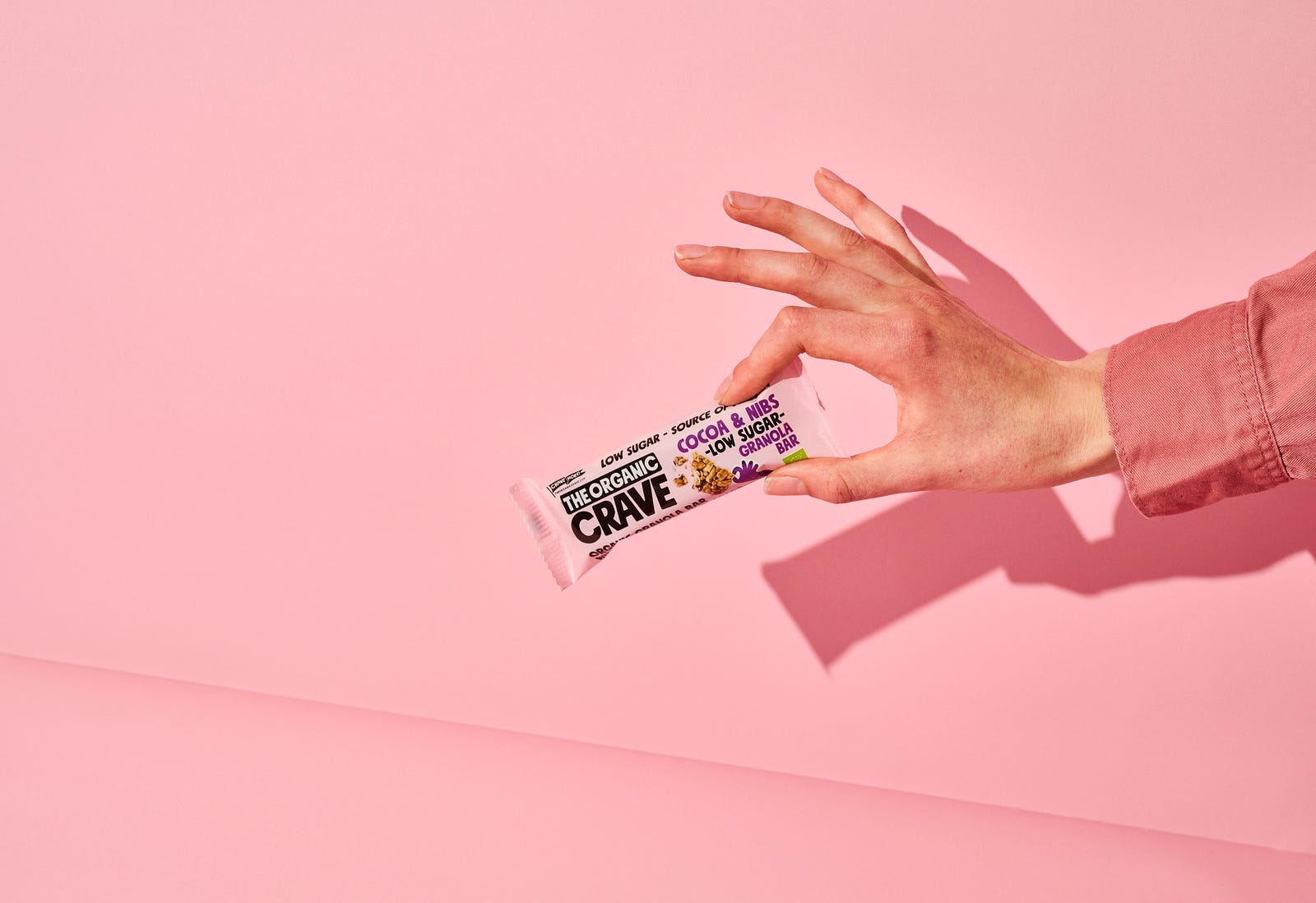 A hand emerges from the right to pinch (with thumb and index finger) a protein bar. Pink background. Choose a bar that meets your diet aims, but you shouldn’t use protein bars as your main nutrition source.
