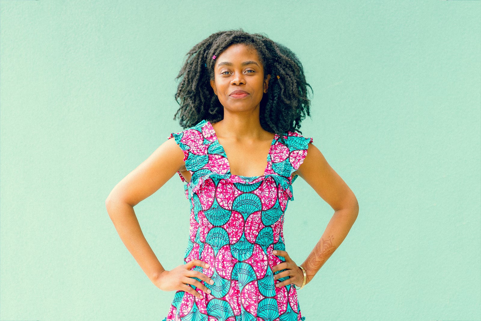 A young black woman faces us, hands on waist. She wears a pink and blue dress with geometric patterns. Light green background. Colorectal incidence and mortality is rising among young adults.