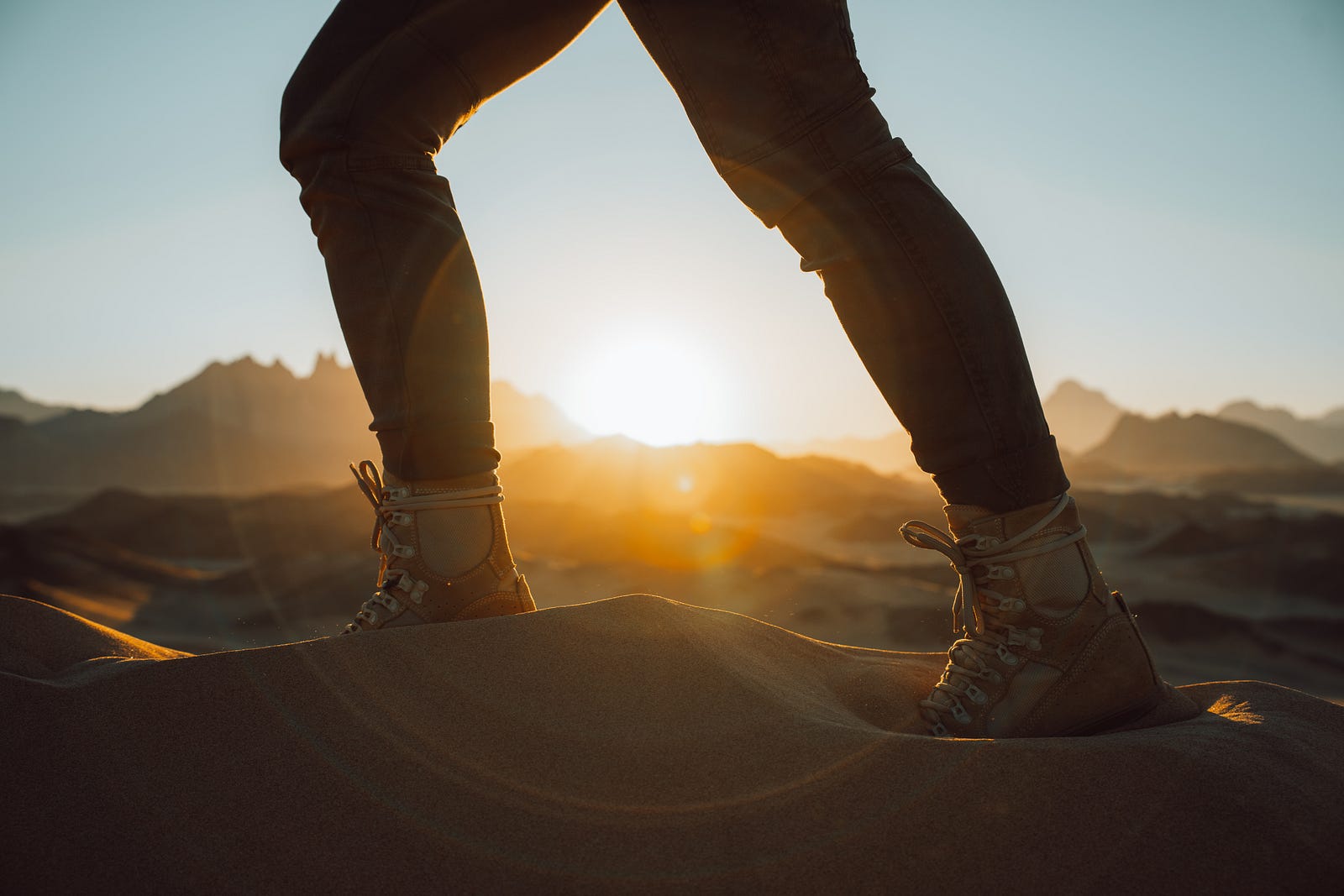 We see a person (from mid-thigh down) walking on sand at sunsrise. Incorporating some bodyweight activities can lead to a more challenging workout. Alternative terrains (such as a beach) can introduce more challenges, too.