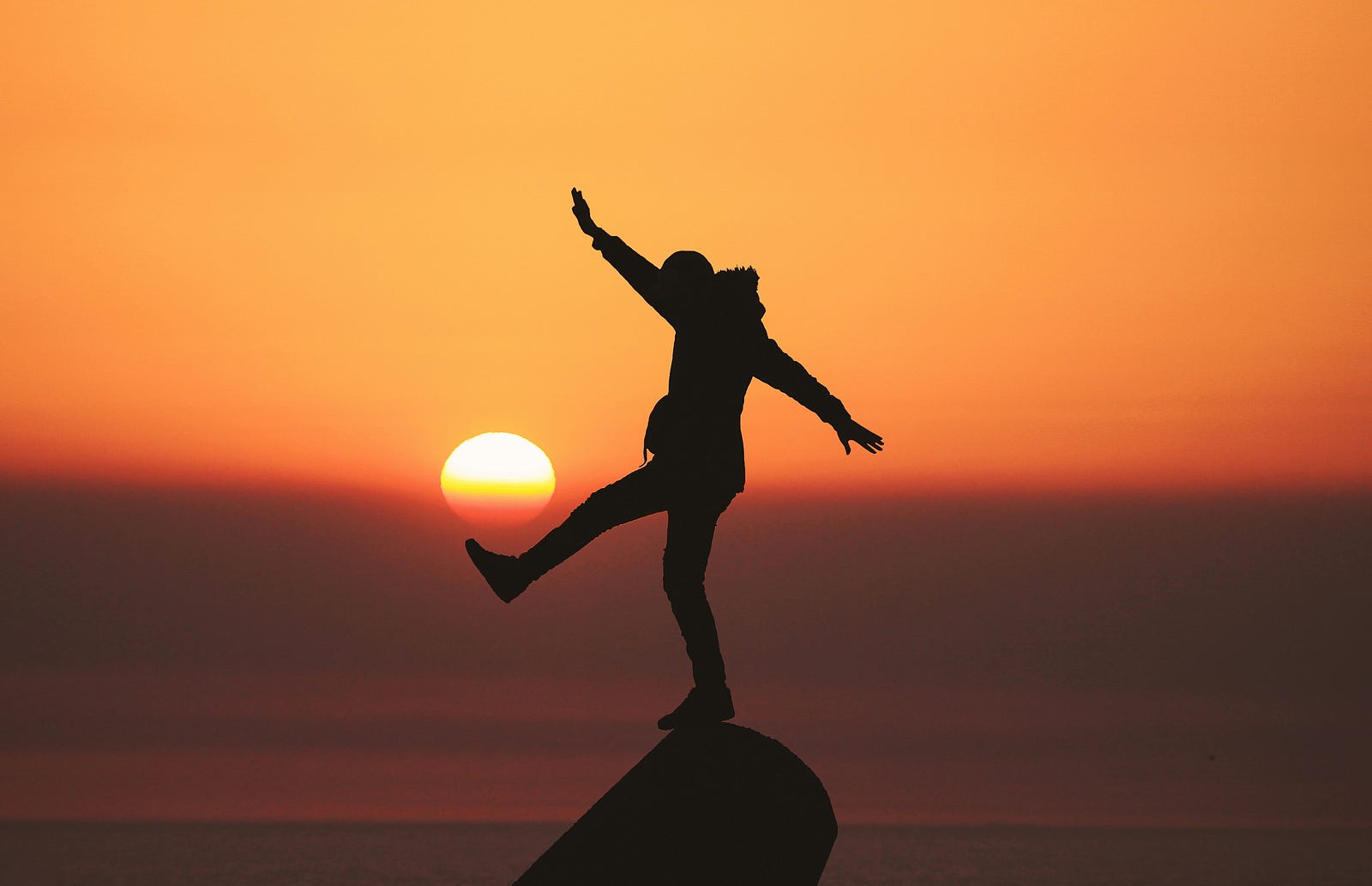 A person balances on a rock as the sun sets in the distance.