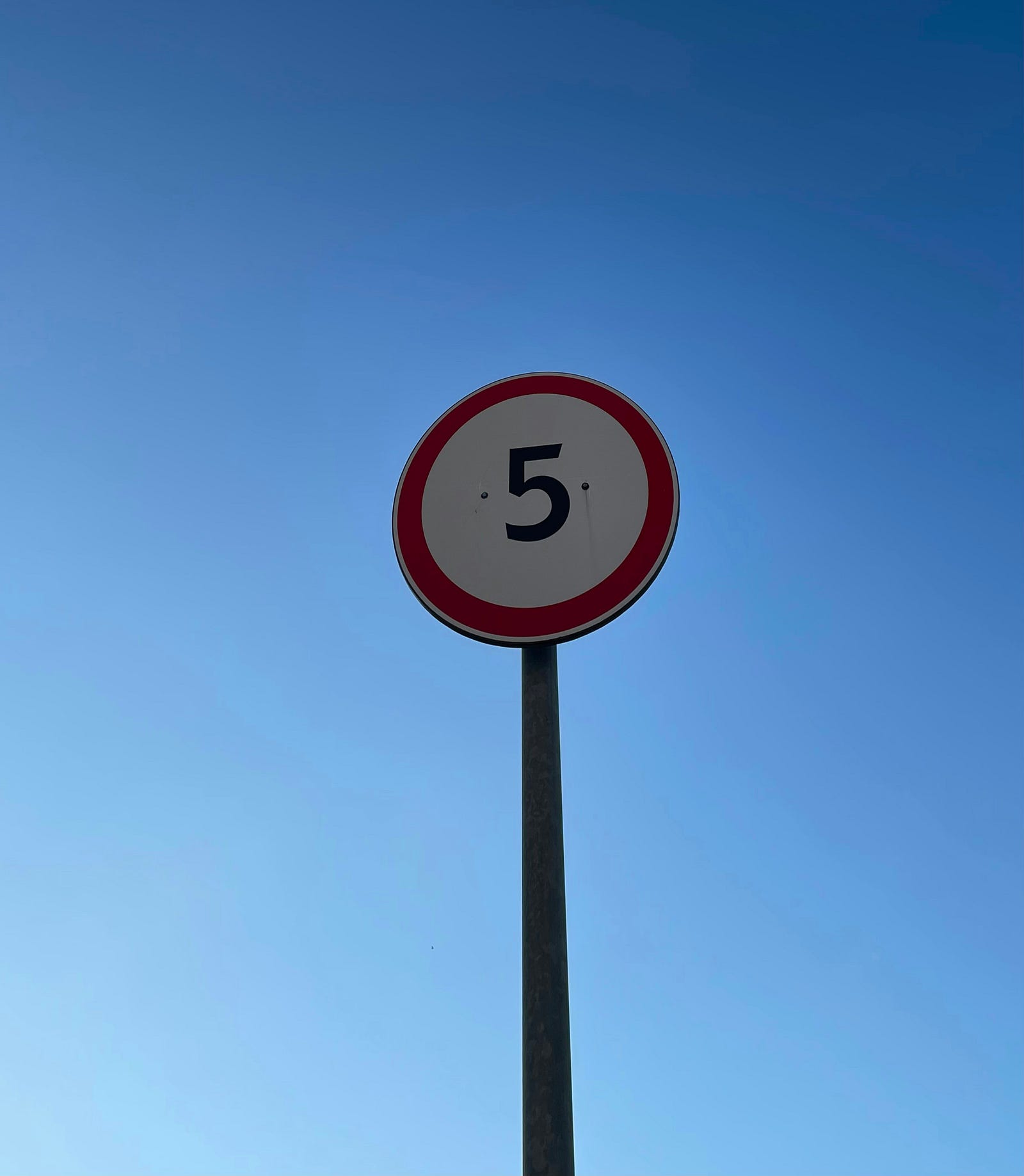 A round sign (on a narrow long silver pole) has the number five in black. The number is surrounder by a red-rimmed white area.