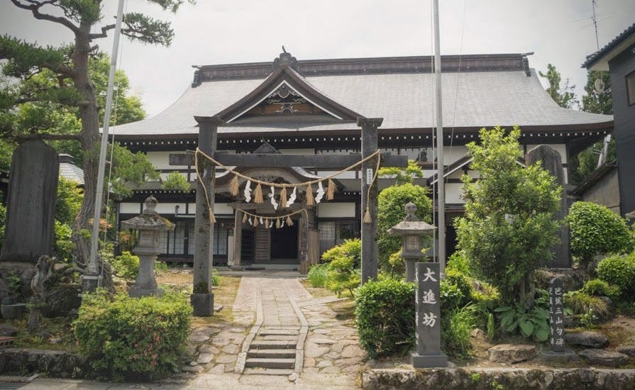 The grand building of Shukubo Daishinbo pilgrim’s lodge on Haguro-san. In the front is the ceremonial Torii gates decorated with a straw rope with white lightening bolt-shaped tassles. Monument to Matsuo Basho visible to the right.
