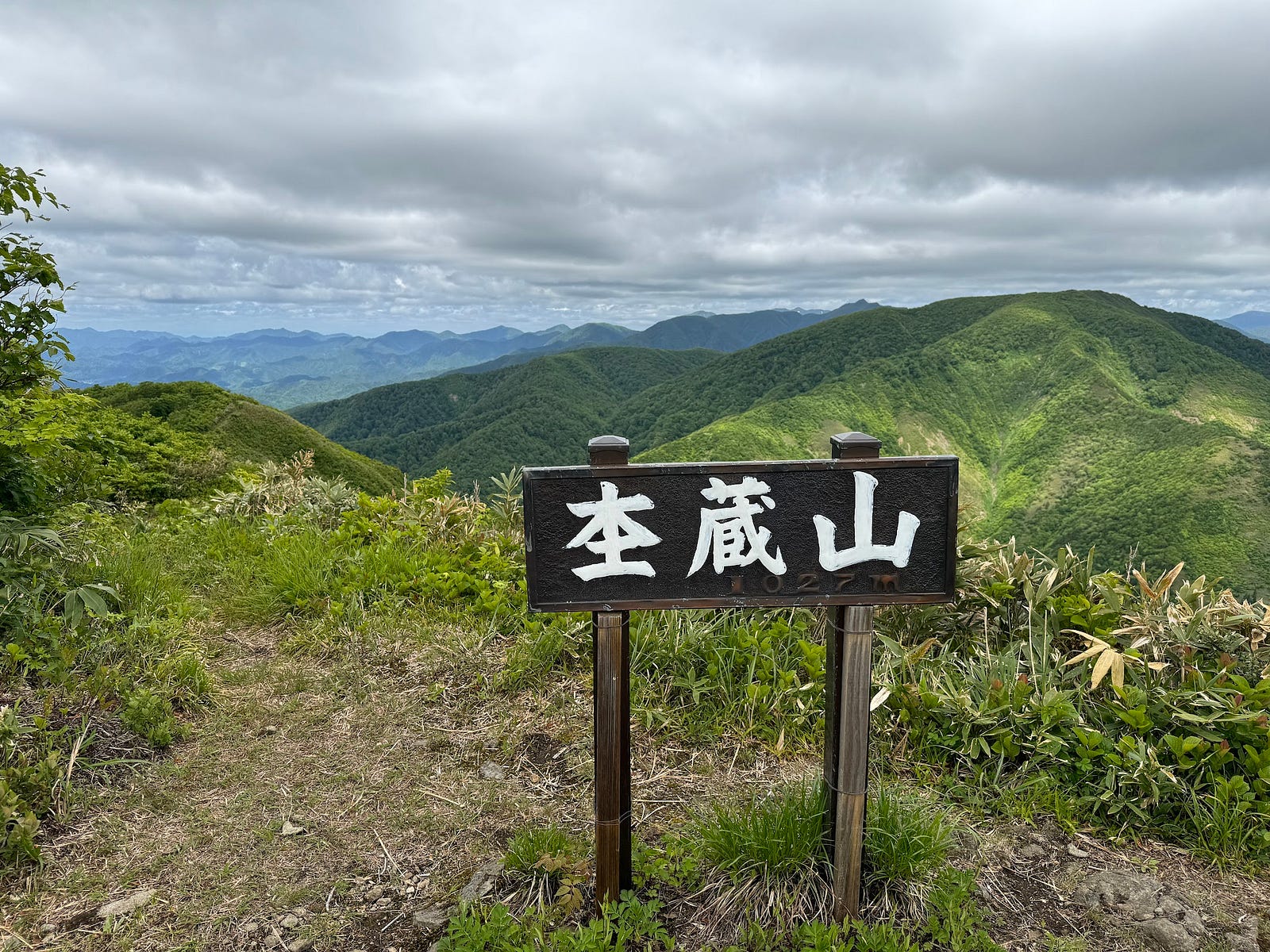 A sign at the summit of Mokuzo-yama with the green Kamuro Renpo (alps) under a deep grey sky.