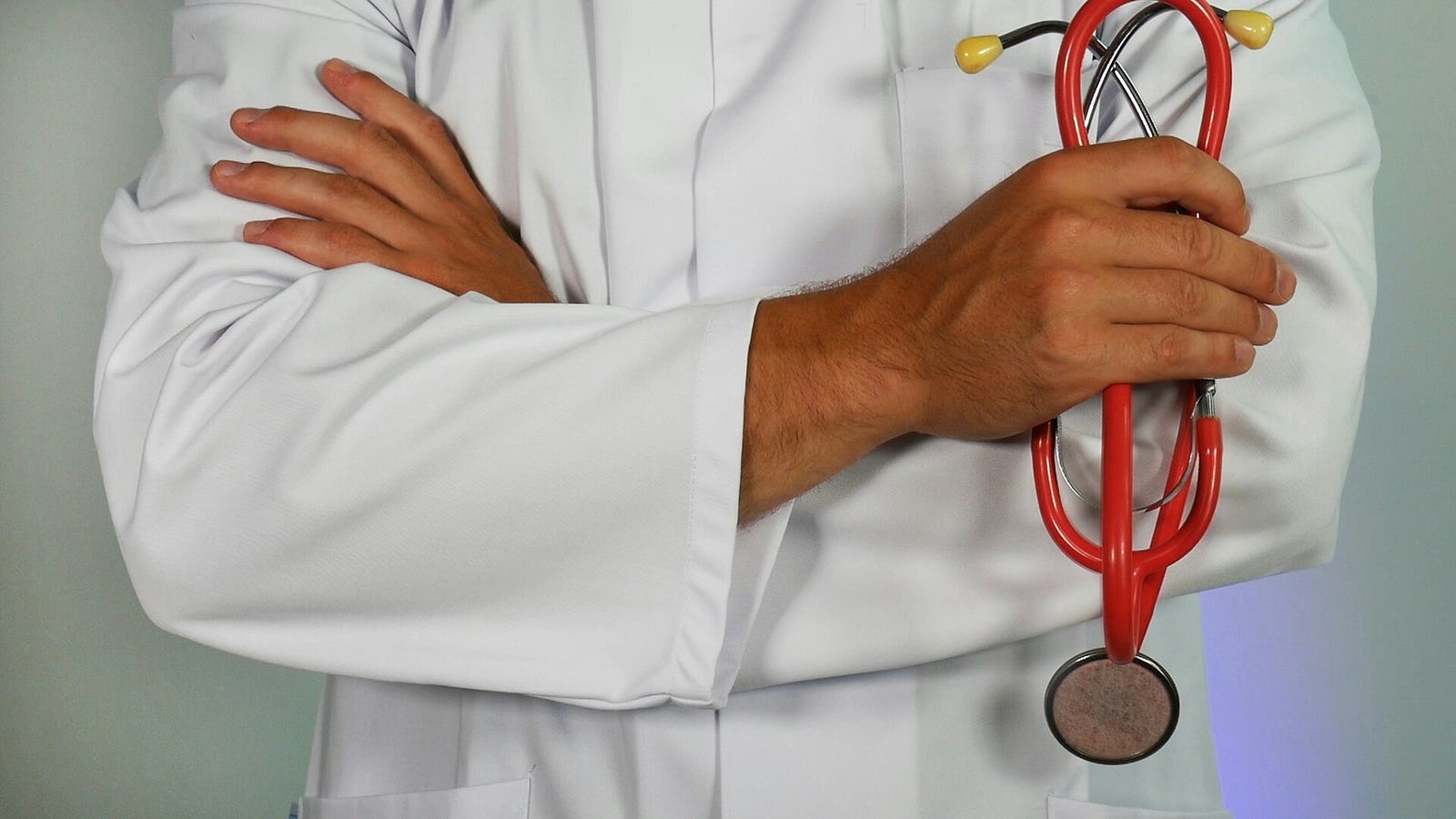 A doctor in a white coat stands with their arms crossed and a red stethoscope in their left hand.