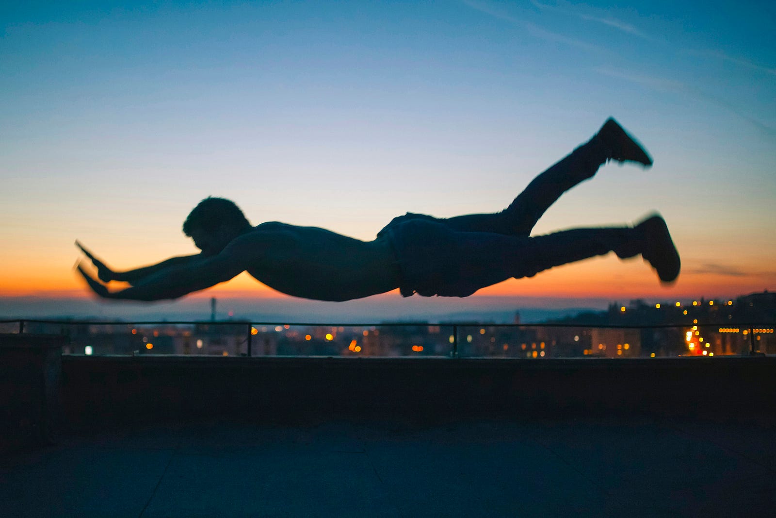 A man floats, in the superman pose, with a city in the background at sunset.