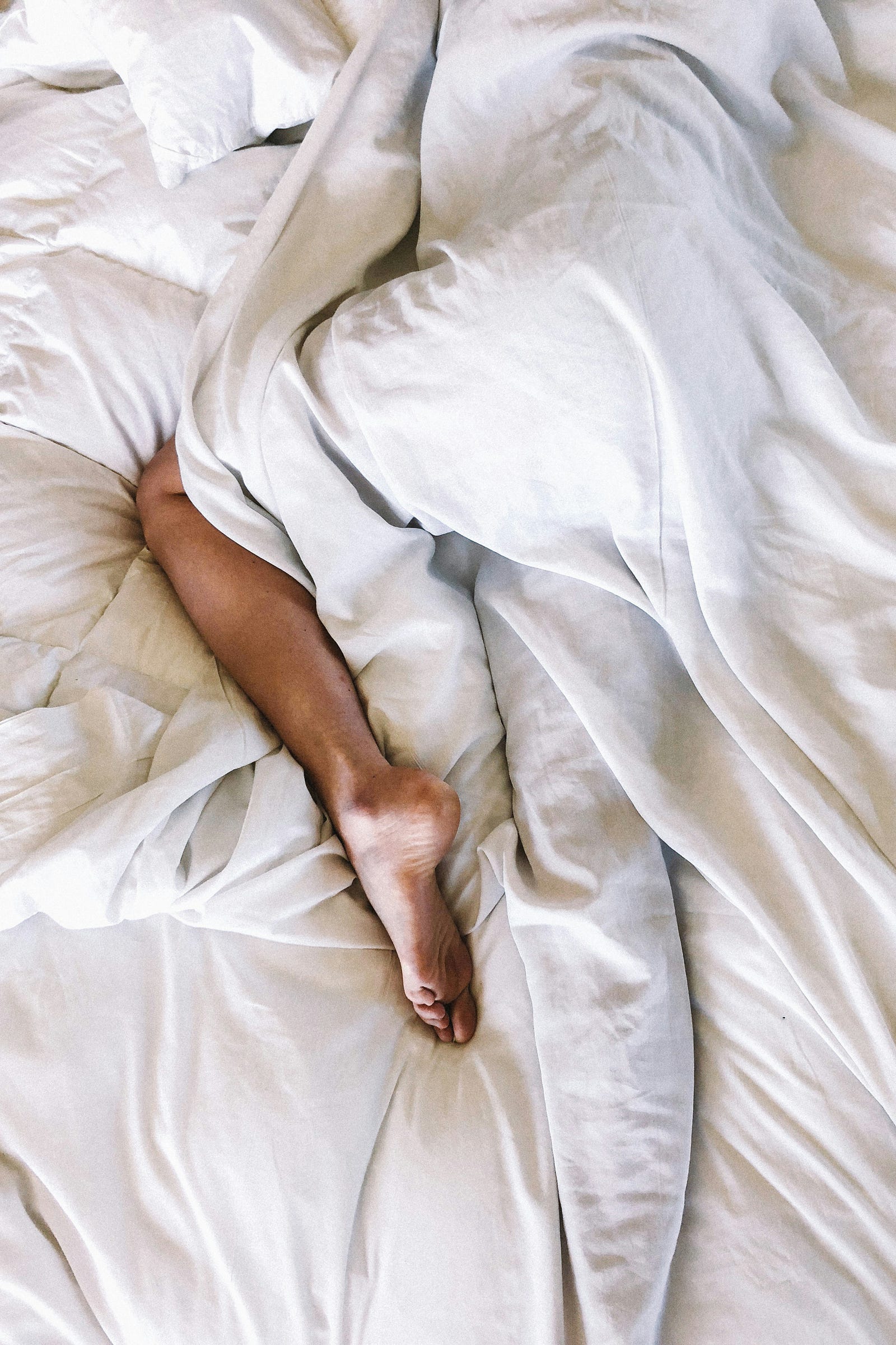 A person’s leg peeks out from white bedding. Poor sleep can lower testosterone hormone levels.