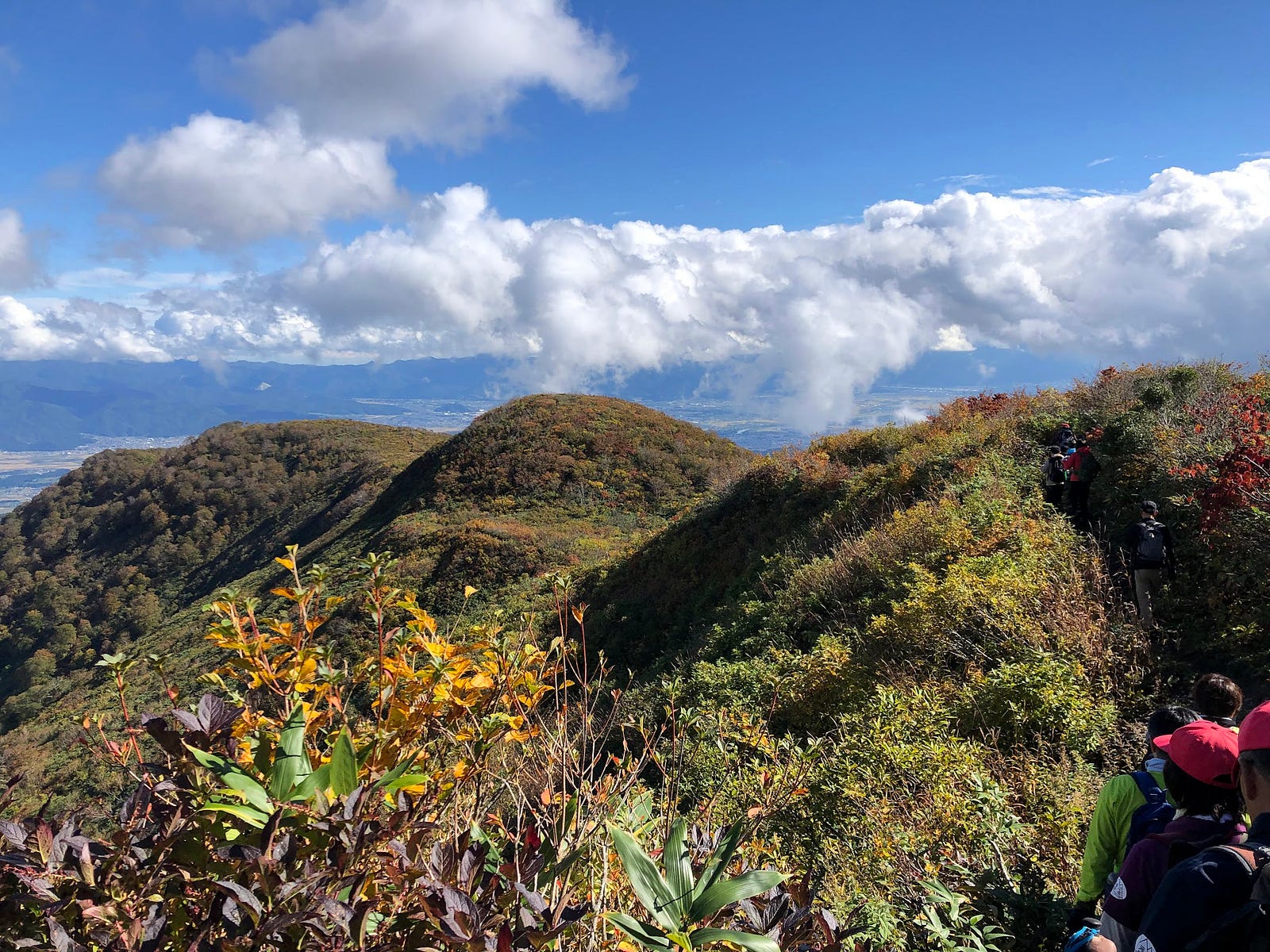 Hikers stare down to the bumpy peaks along the ridge of the extinct volcano that makes up Murayama Ha-yama, blue sky and clouds in the background