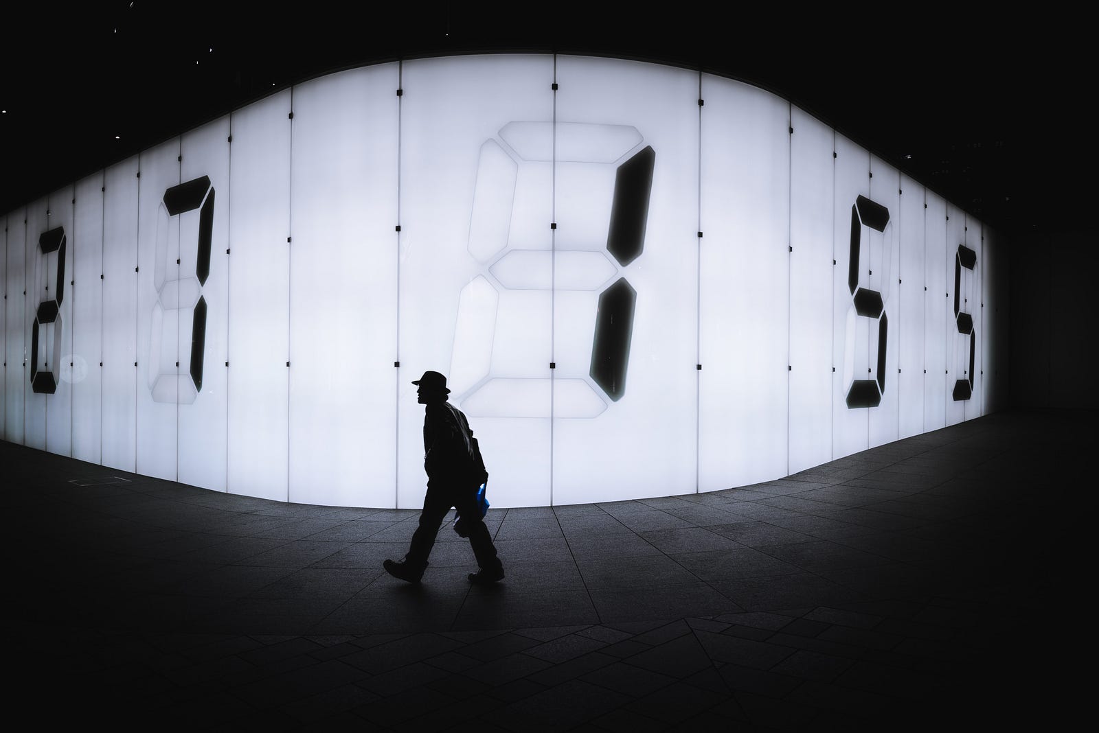 A black and white image of a solitary figure walking from right to left. There is a wall of numbers (white and backlit) in the background. A new study shows that, for cardiovascular disease, walking daily at least 2,337 steps reduced the risk, with each extra 500 daily steps associated with an additional seven percent relative reduction in risk.