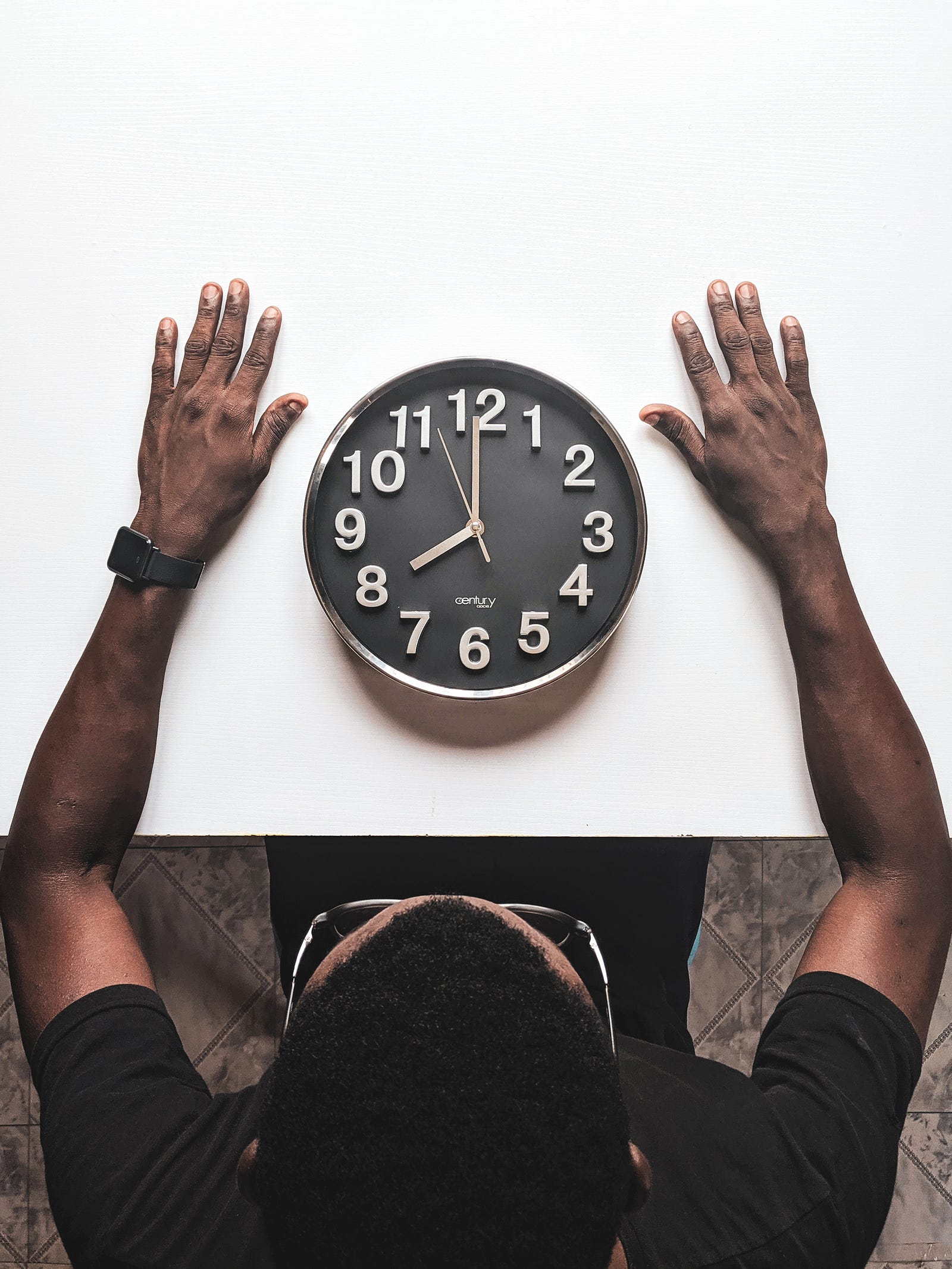 A clock with a black face *and white numbers and hands) sits on a white table. A person extends their arms around the clock. Why might vaccination effectiveness be linked to the time of day? Researchers think that the circadian rhythm — 24-hour cycles that are part of the body’s internal clock, running in the background to carry out essential functions and processes — affects the body’s response to the vaccine.