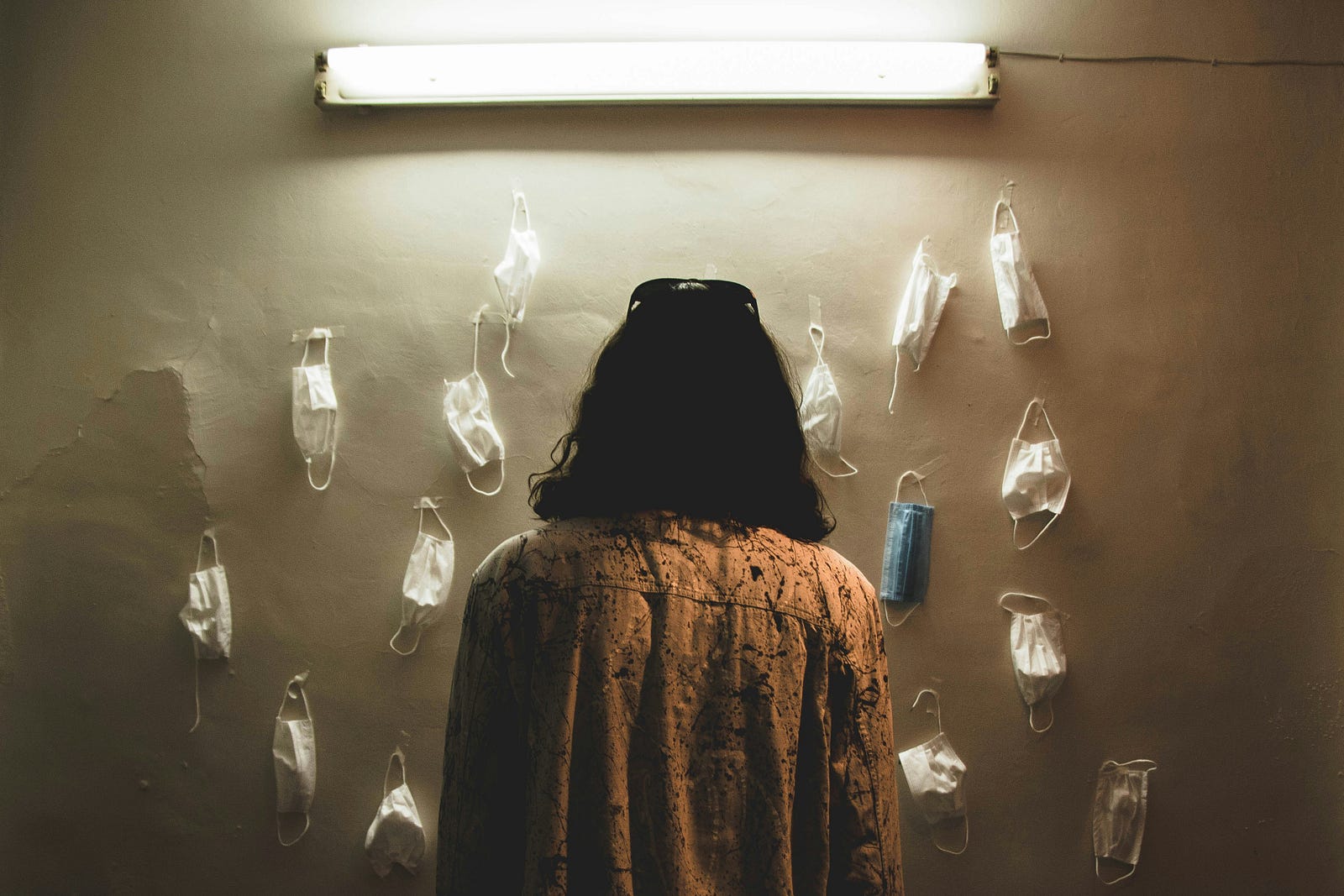 A woman faces away from us, as she looks at a couple dozen surgical masks hanging from a wall.