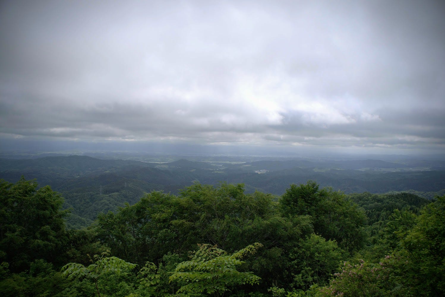 A dramatic sky above and green forest in the foreground with the Shinjo Basin of Yamagata Prefecture in the background.