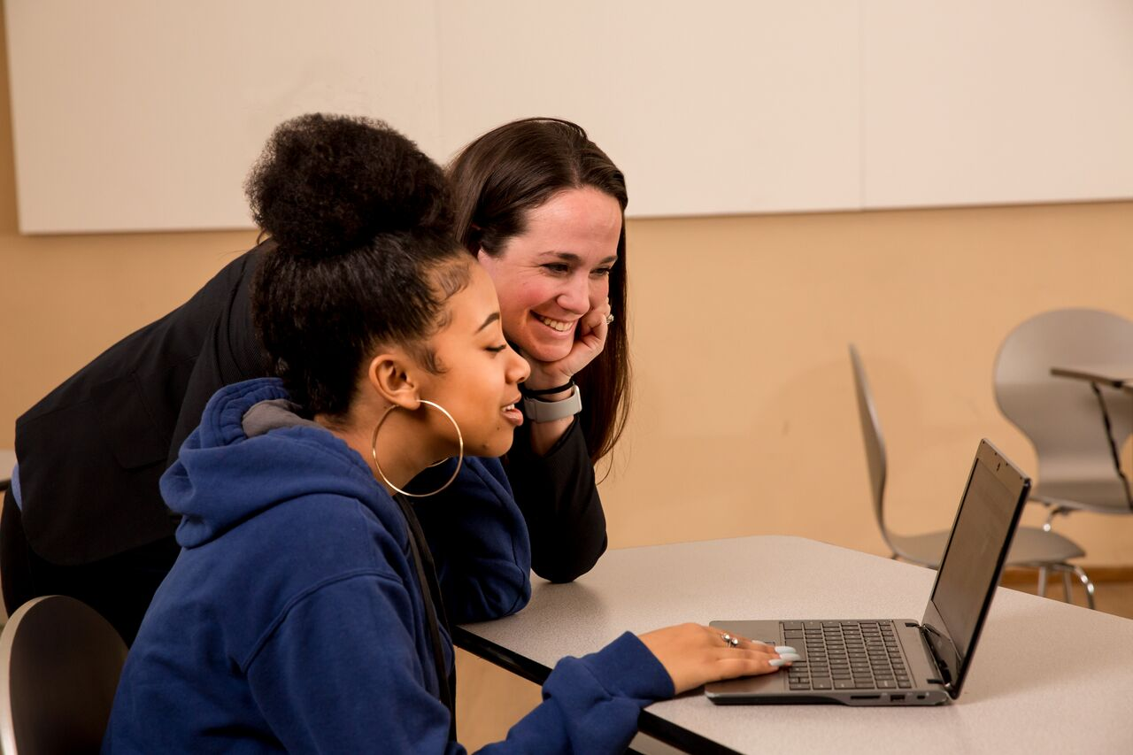 A teacher leaning over a student's shoulder and helping her on her laptop.
