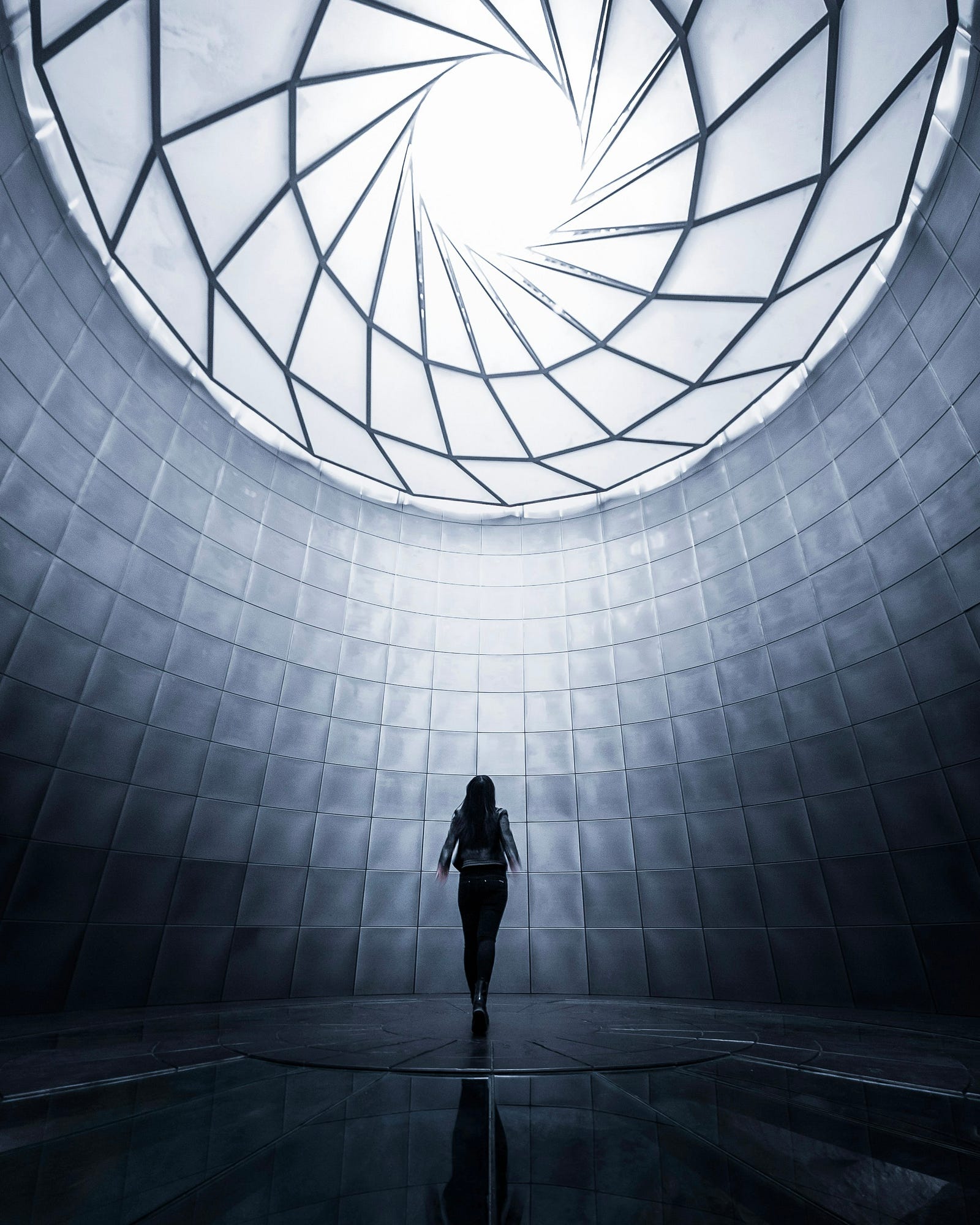 A person walks in a large room with a round glass ceiling insert showing the sky. The image is in black and white.