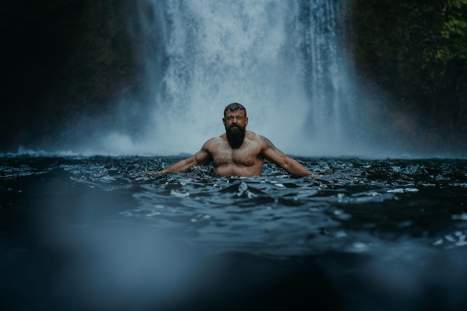 A muscular white man stands in the water in front of a waterfall. Weight training helps build and maintain lean muscle mass and contributes to bone density, which is particularly crucial as we age.
