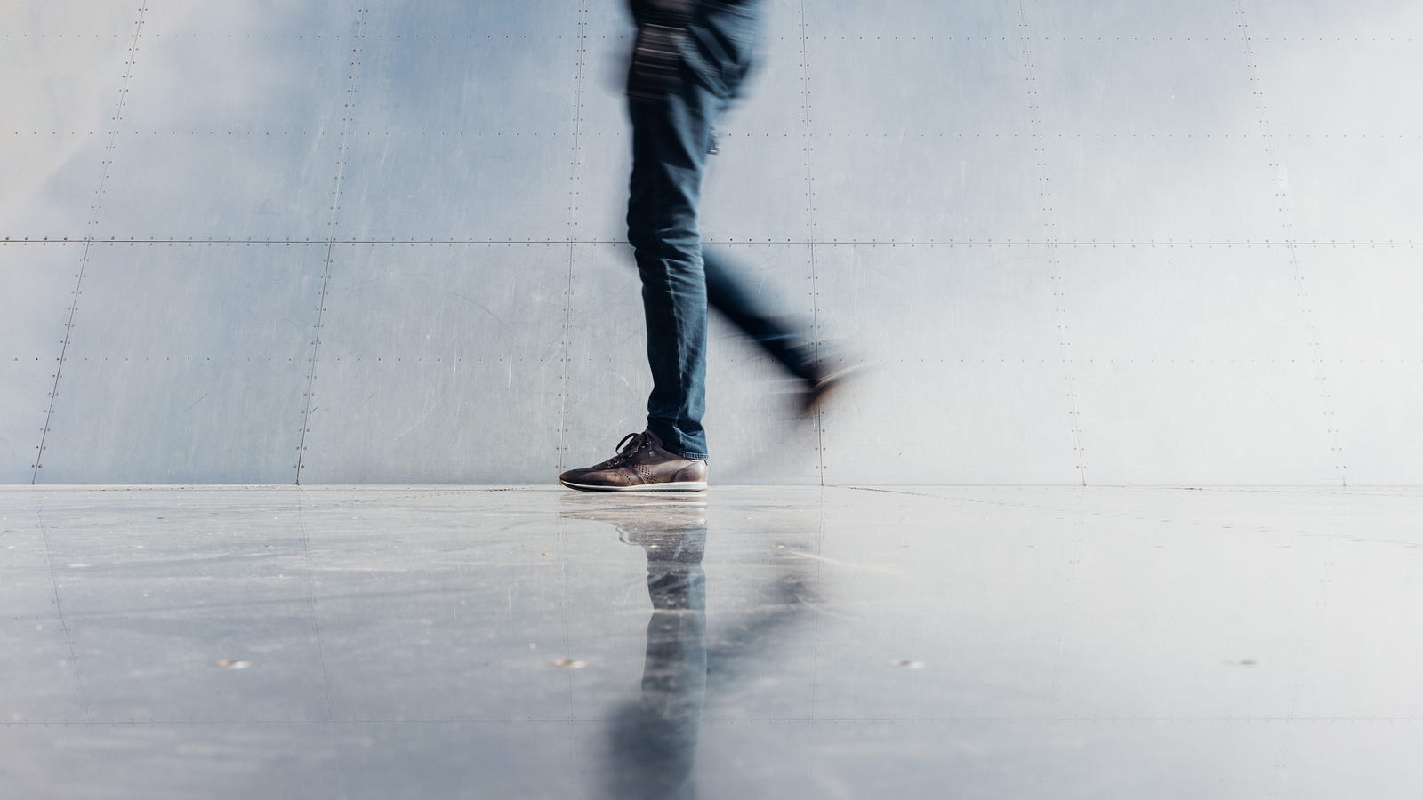 We see a person (from mid-abdomen level down) walking from right to left. S/he wears jeans. The image is slightly blurred, highlighting the movement. There is a building wall (bluish-white) in the background. Walking (or any physical activity) can lower colorectal cancer risk.