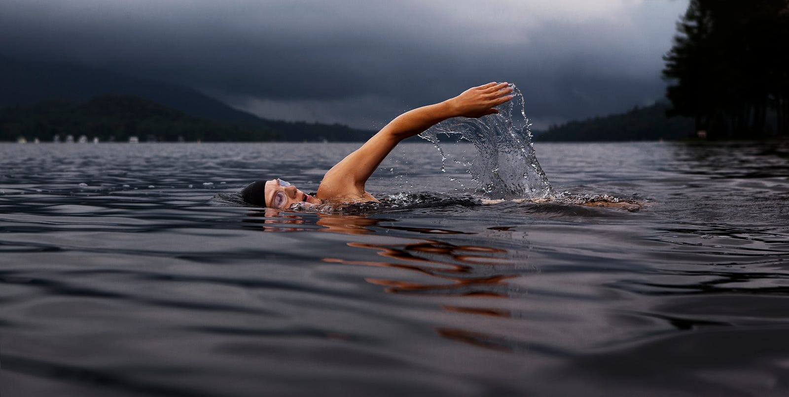 A person swims in a mountain lake from right to left (facing us). Their left arm is raised above the water. Swimming is an exercise that can boost your brain.