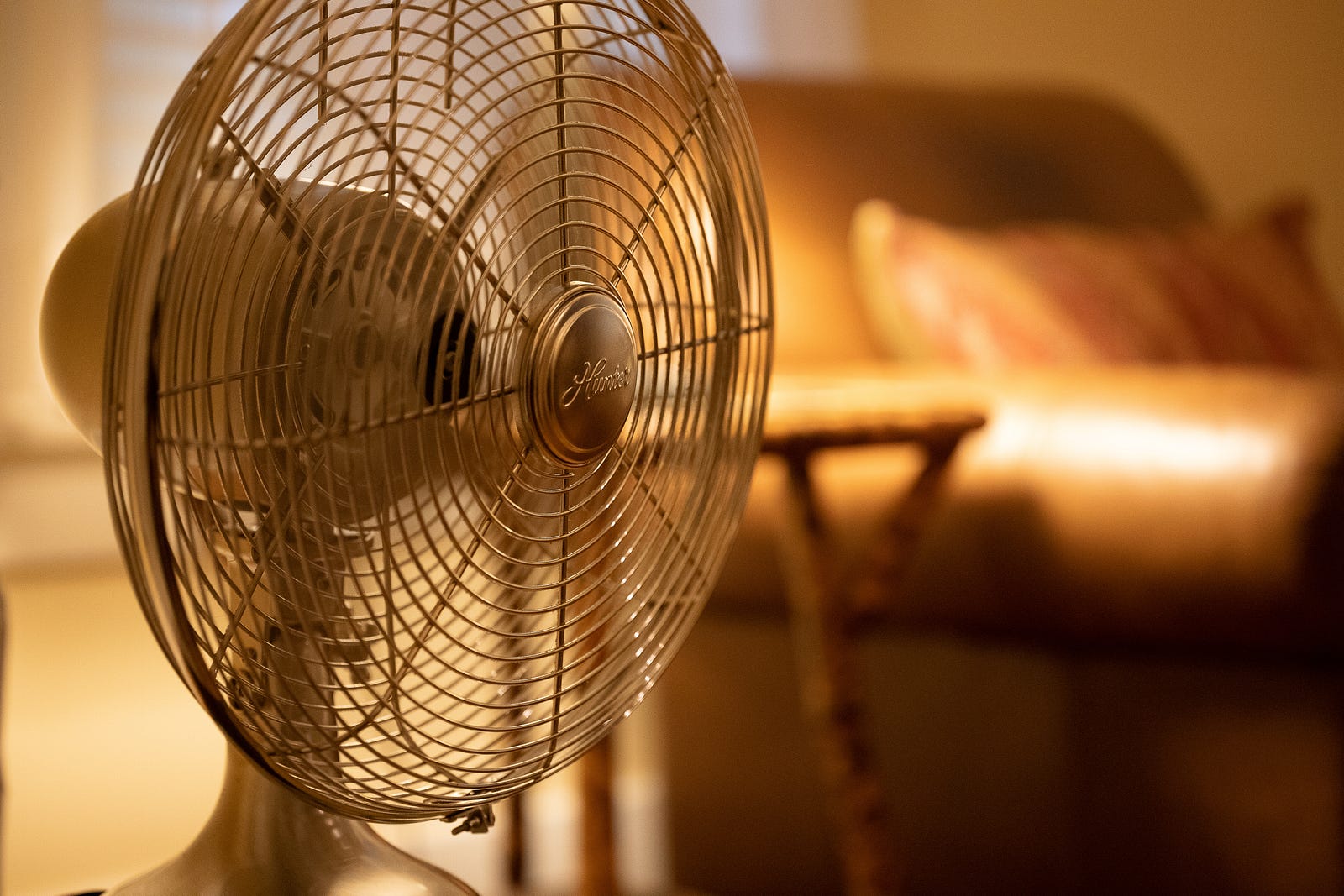 A sepia image of a fan in the foreground (with a brown sofa on the right side of the background). Using a fan or air conditioner in your bedroom can create a cool and comfortable environment for sleeping. These appliances help circulate air and lower the ambient temperature, promoting better sleep quality.
