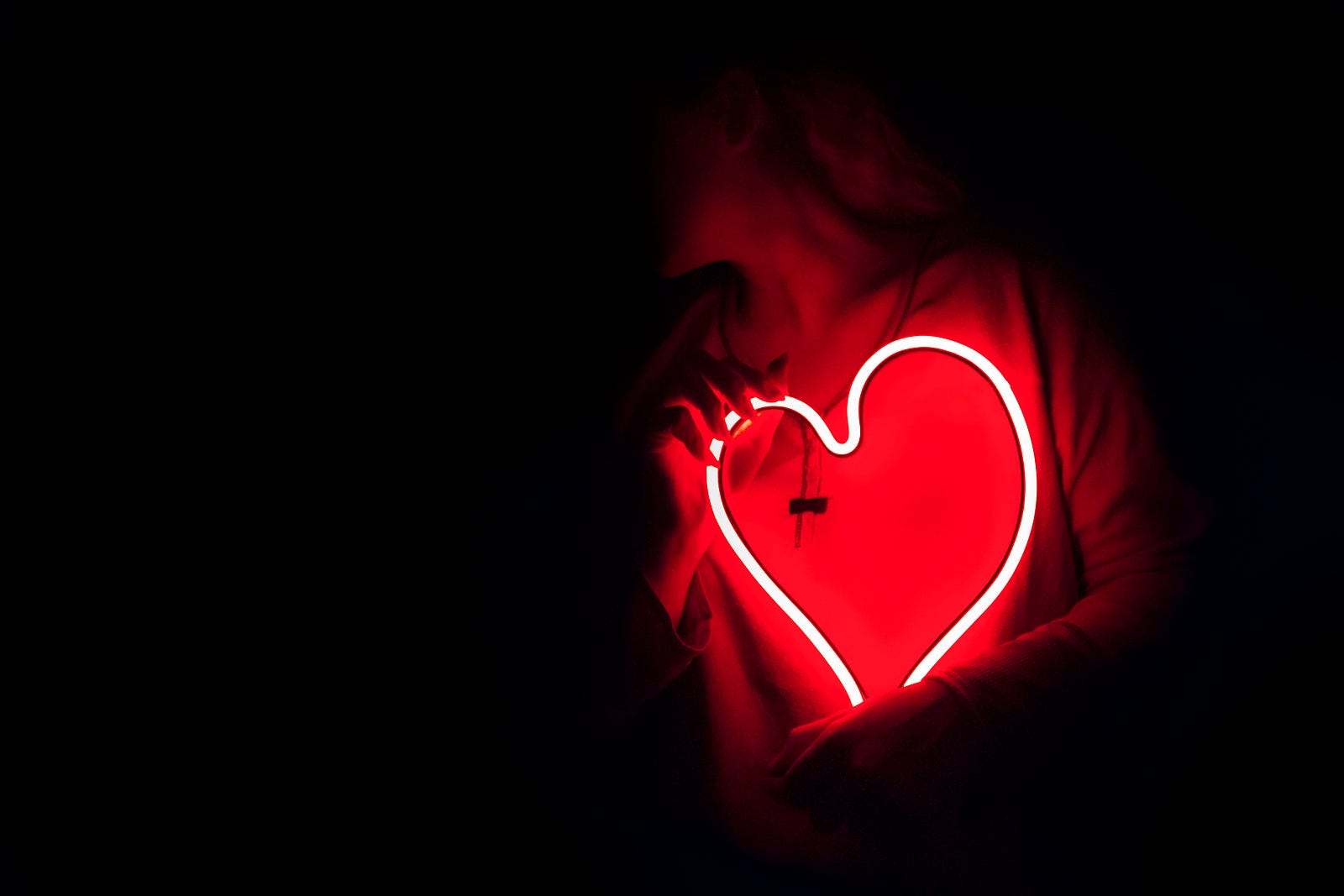 A woman clutches (against her left chest) a heart made of neon.