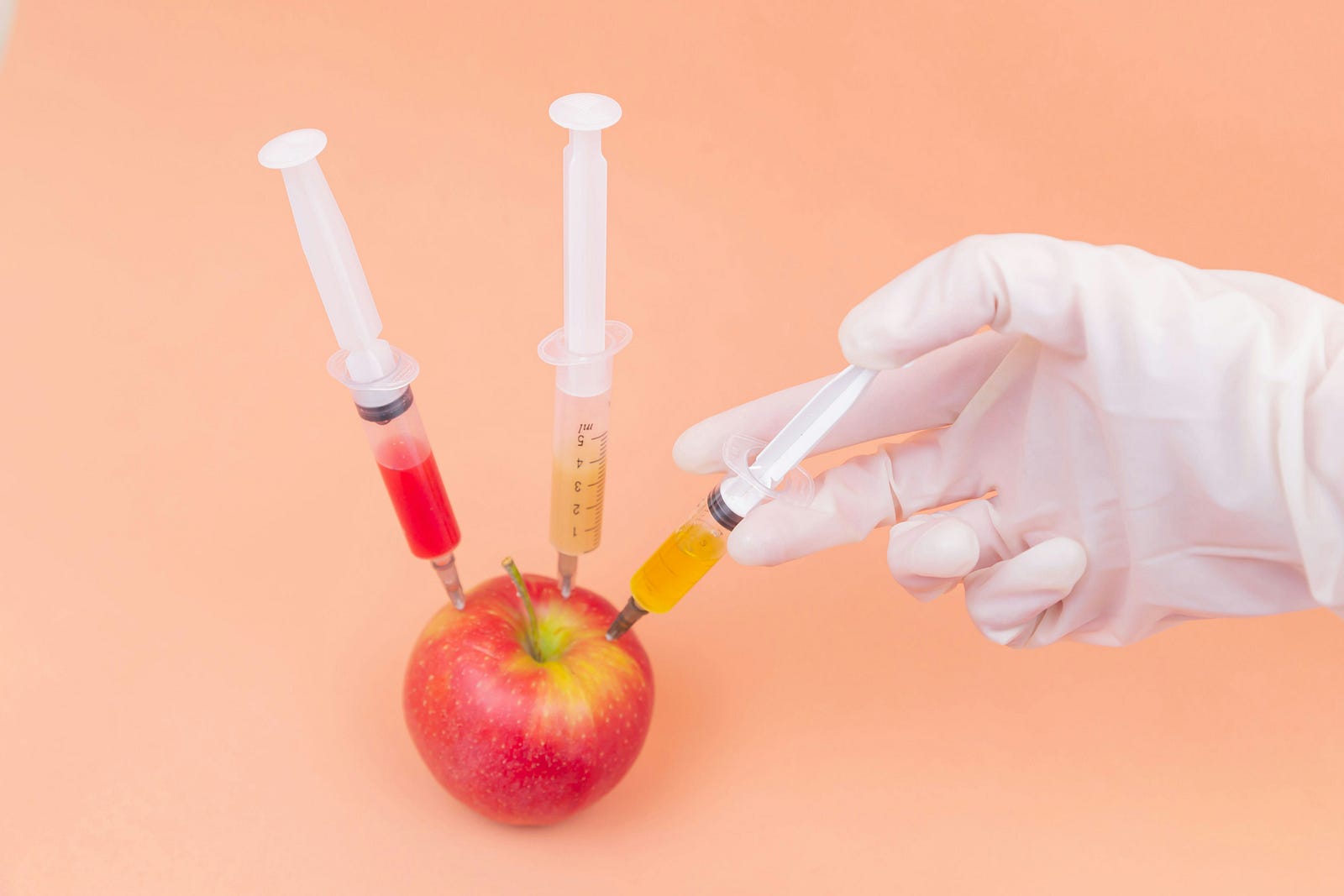 An apple with three syringes (with various colored additives) sticking into it.