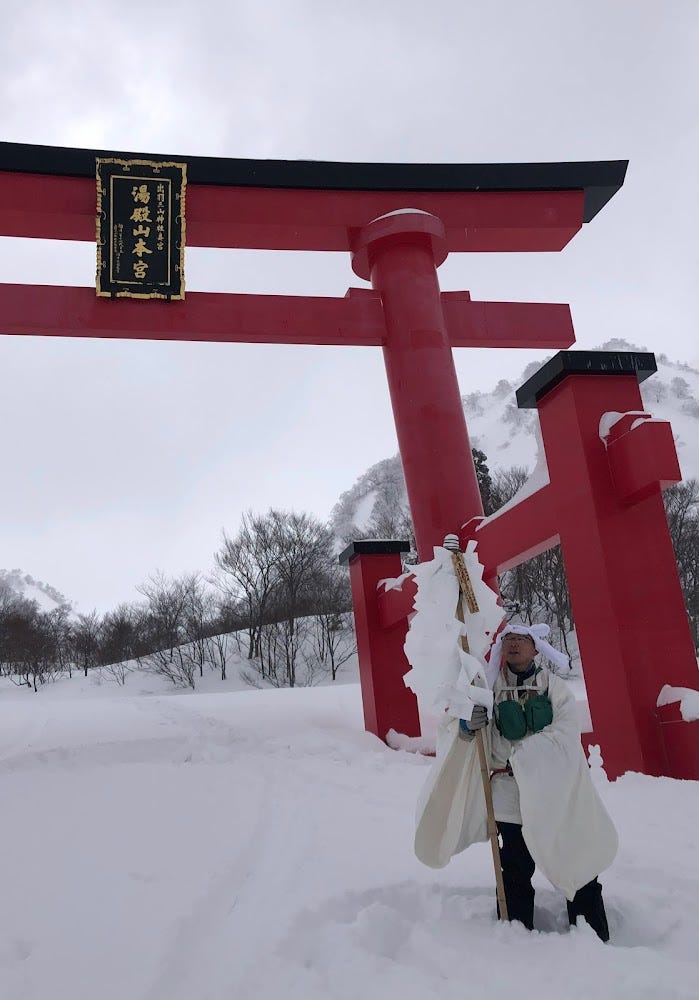 A yamabushi holding a bonten stands beneath the shrine gates of Mt. Yudono in the middle of winter