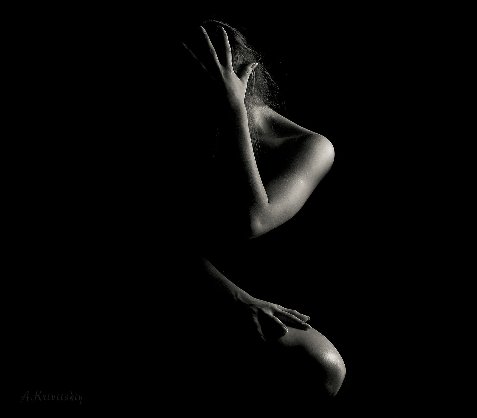 A nude woman faces us with her right arm covering her face . The image is dark, but we see her left side highlighted by light.