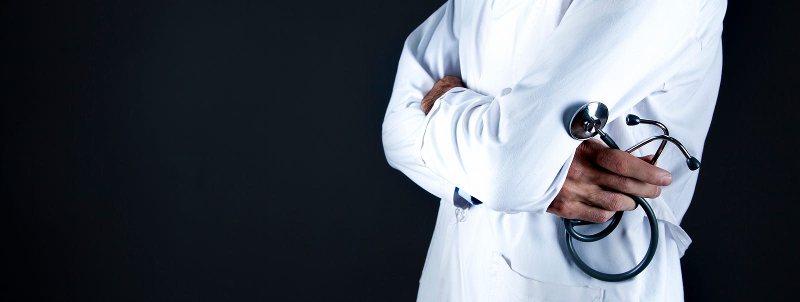 We see a doctor (from waist to upper chest) in profile at the right of the image, wearing a white coat with arms crossed. The right hand holds a stethoscope. According to the manufacturer, the most common side effects of Veozah include abdominal pain (4.3 percent versus 2.1 percent for placebo), diarrhea (3.9 versus 2.6 percent), insomnia (3.9 versus 1.8 percent), and back pain (3 versus 2.1 percent).