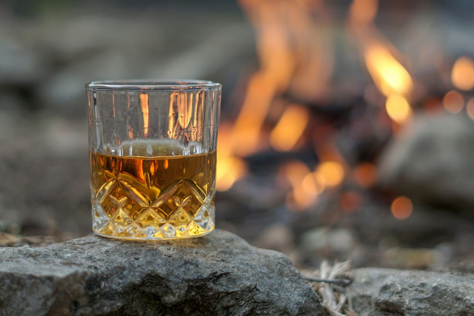 A short glass of alcohol sits on a piece of lava. There is fire burning in the background.