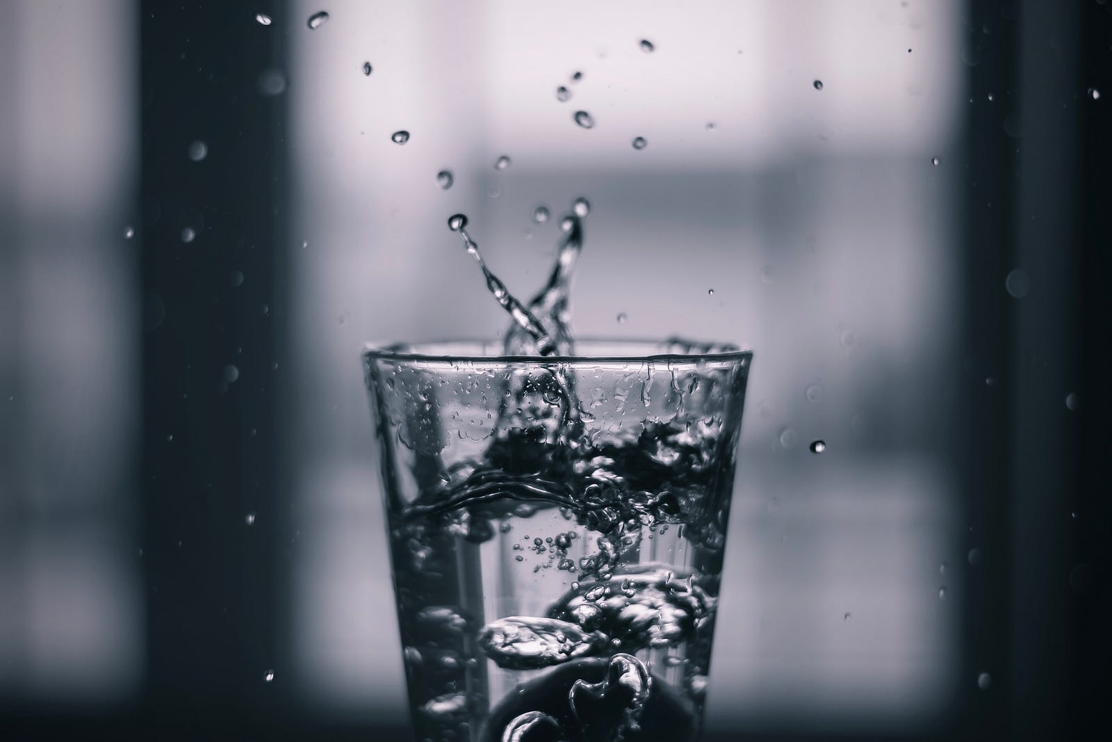 Water drops from above into a clear medium-sized glass, with water splashing upwards out of the glass.