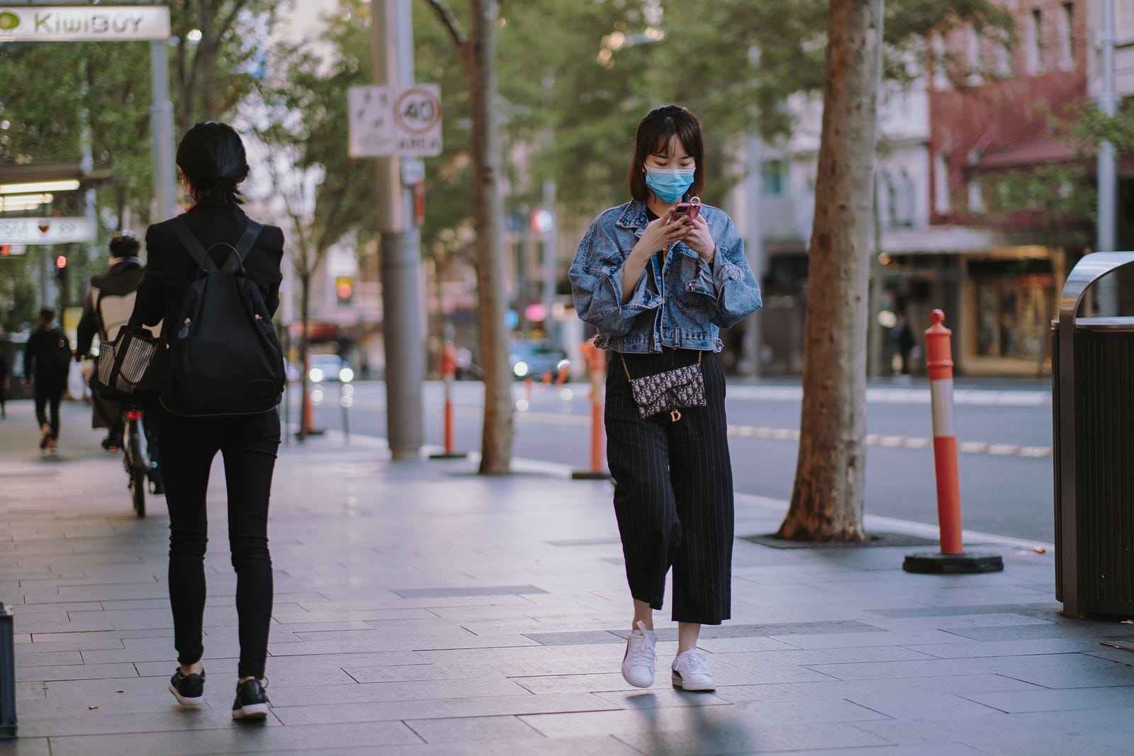 A few Asian individuals walk on a sidewalk. One wears a face mask as she manipulates her smartphone. A new study shows that a COVID infection provides considerable protection against future severe infection.
