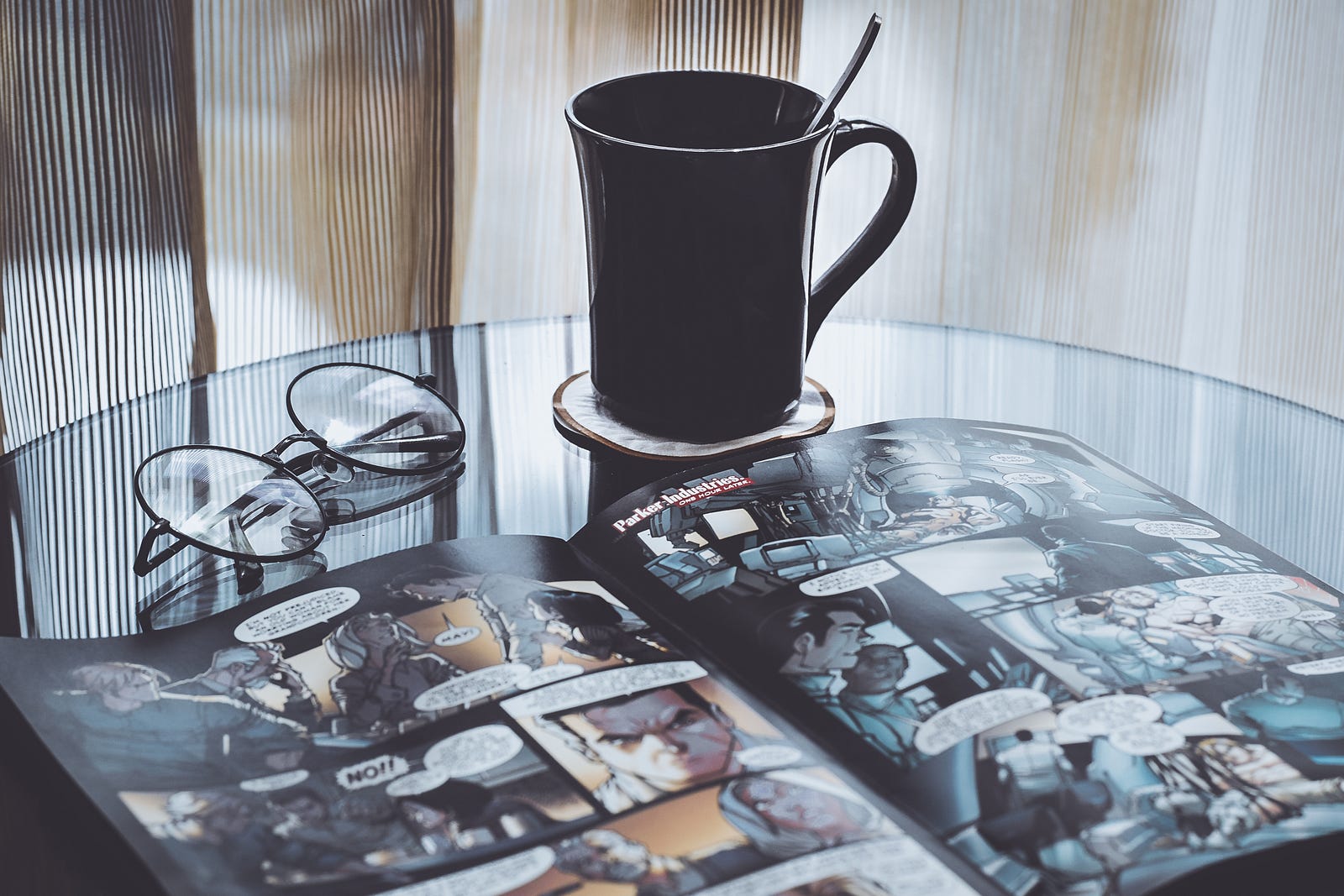 A photo of glasses, a mug of coffee, and a comic book and a table.