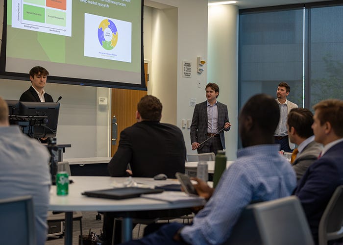 A group of individuals standing at the front of a classroom with two large projection screens, presenting to an audience of seated attendees.