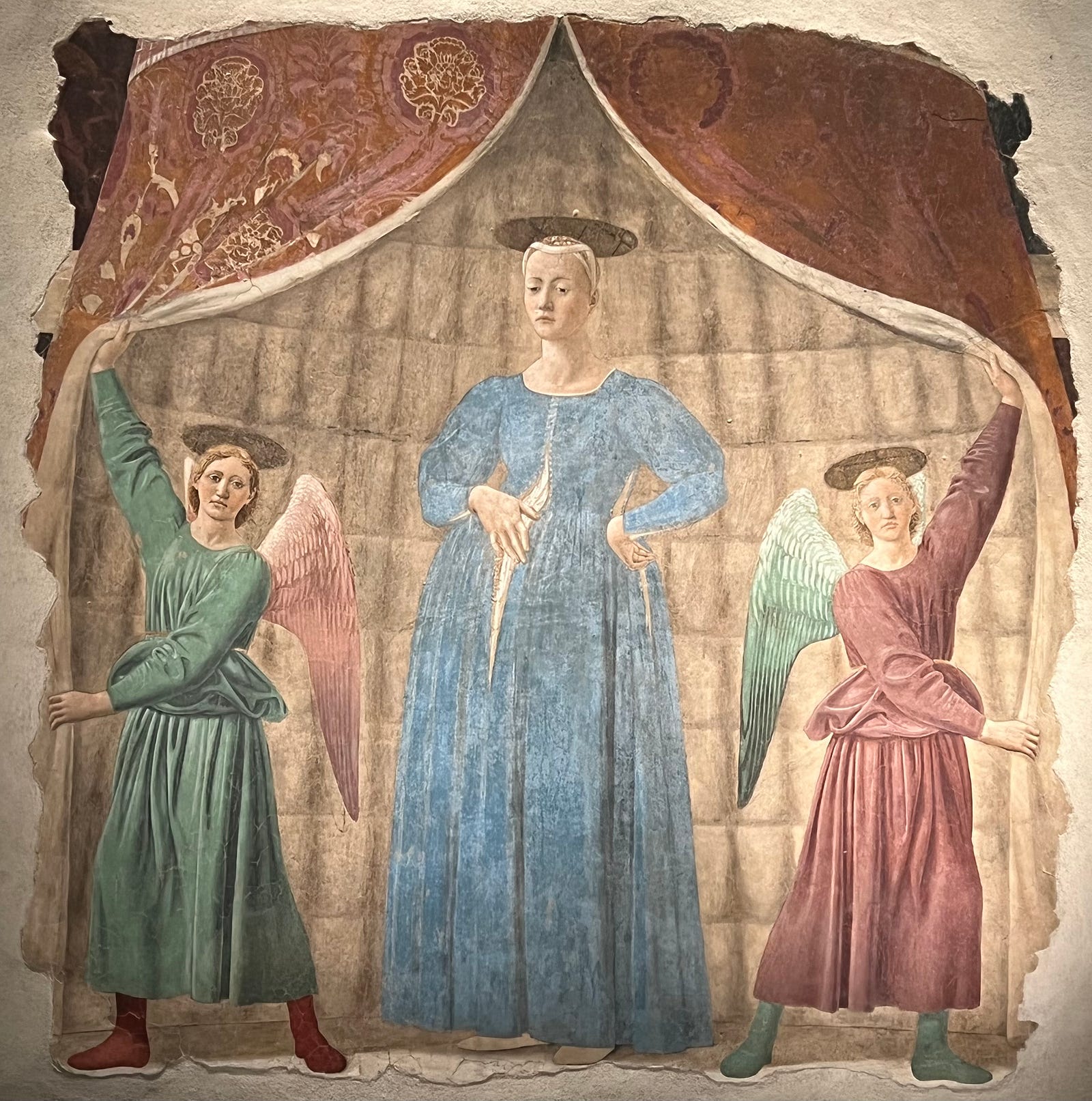 Photo of the fresco Madonna del Parto of Monterchi by Piero della Francesca (c 1460), showing a very pregant mary in blue frock, stading within a tented encousure whose flaps are held open by a pair of angels who look directly into the viewer’s eyes.