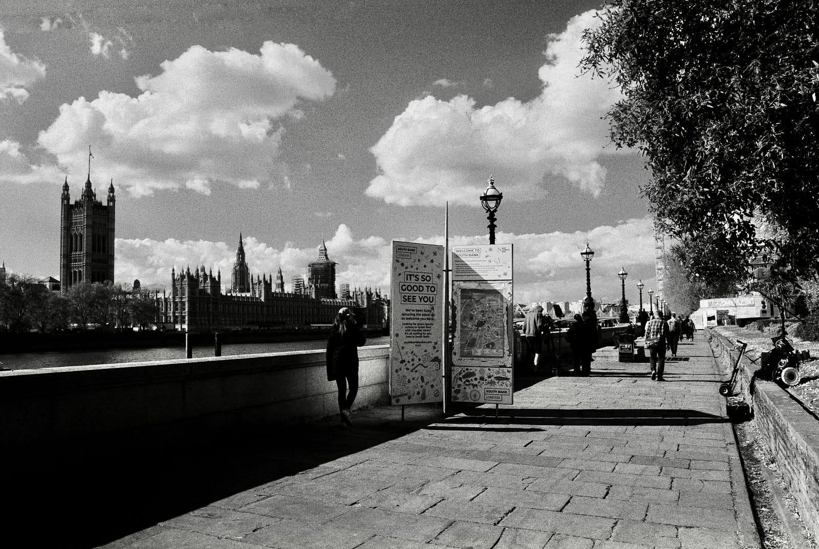 A black and white image of the South Bank at Lambeth, looking towards the Houses of Parliament across the river. People are walking up and down the promenade.