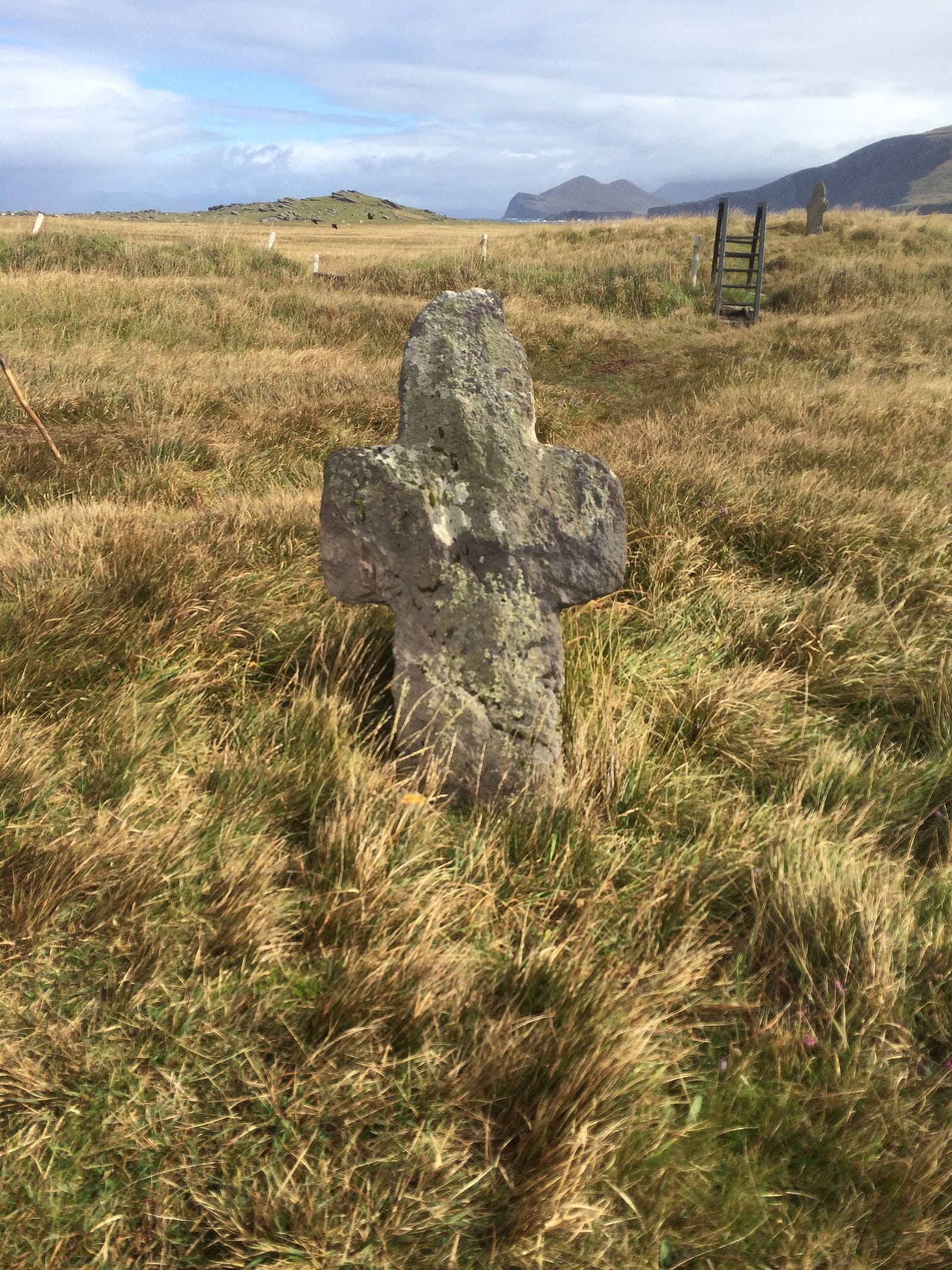 Photo of a standing stone cross at St Brendan’s Well on Valentia Island, Ireland. The cross is rough-hewn and ancient, and stands among the salt grasses.