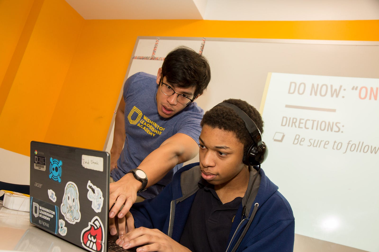 A student working on his computer, with his teacher next to him, pointing at the screen.