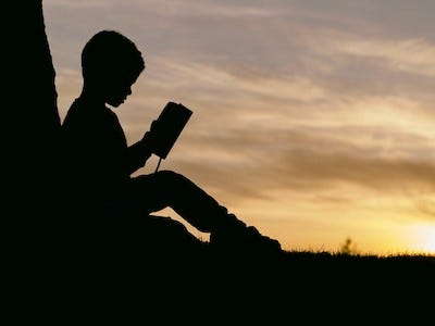 Silhouette of a boy sitting against a tree, reading a book, with the sunset in the background.